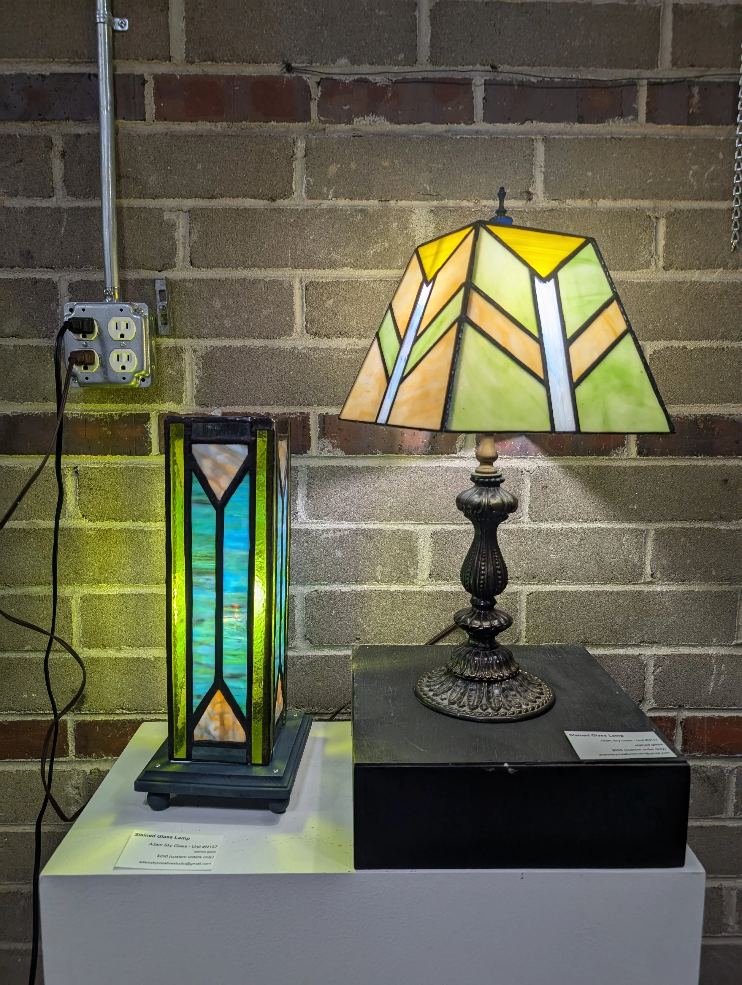 A stained glass lamp with a colorful geometric design on a black base and a Tiffany-style stained glass lamp with triangles and rectangles in yellow, orange, green, and white on a round base.