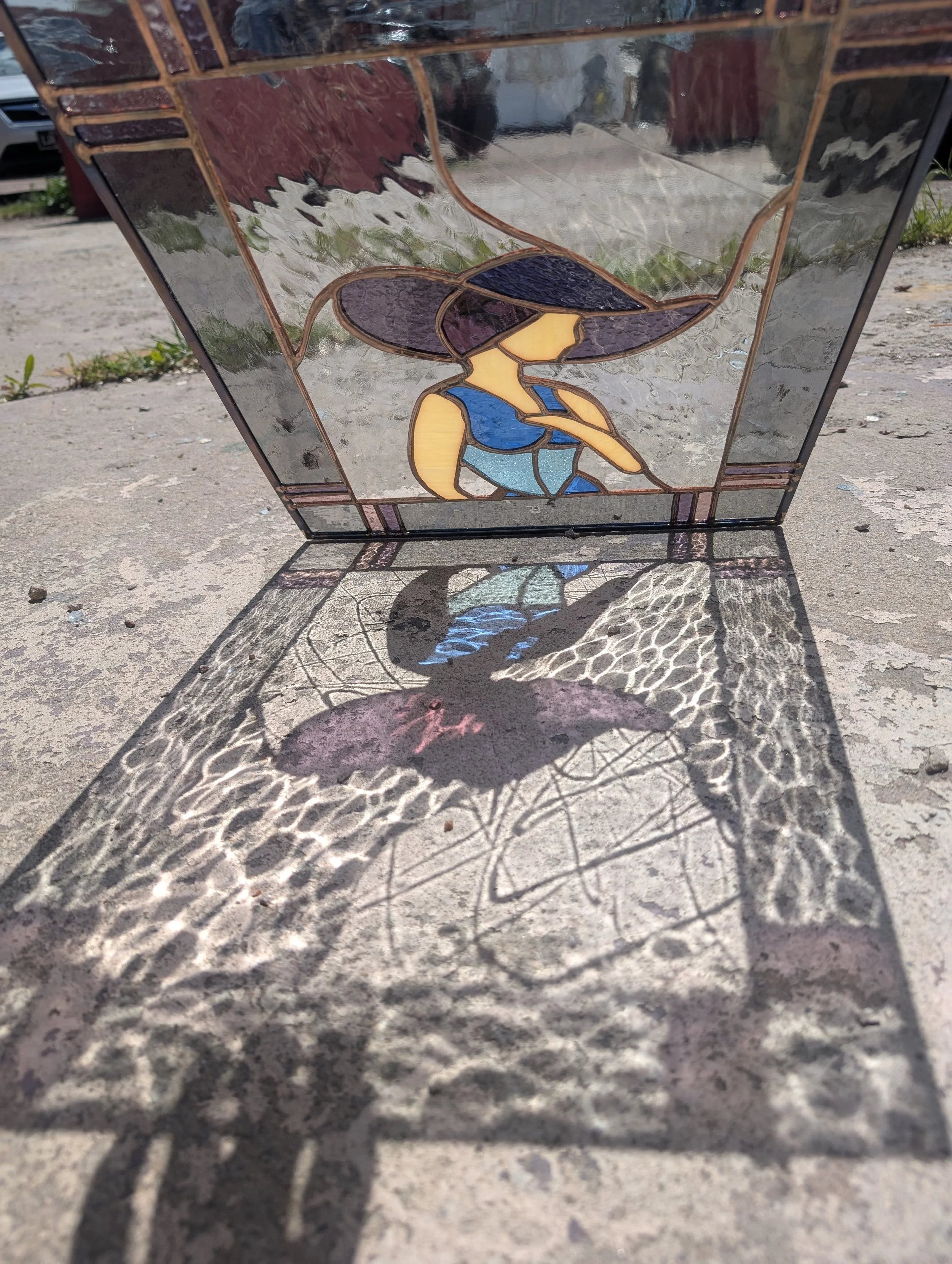 Stained glass lamp with a woman in a blue dress and large hat, casting a colorful shadow pattern on the concrete ground.