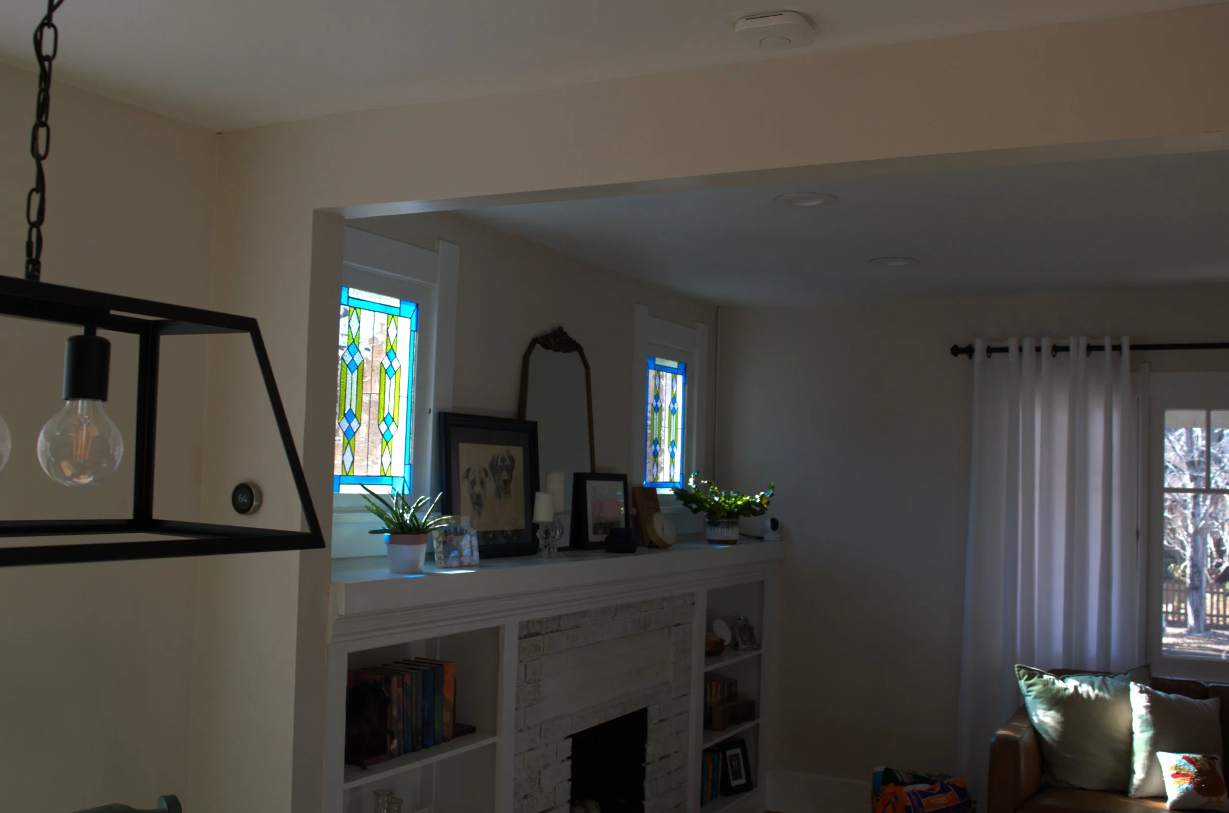 Interior view of a living room with white walls, a fireplace with bookshelves, two stained glass windows, a mirror, framed pictures, plants, and white curtains.