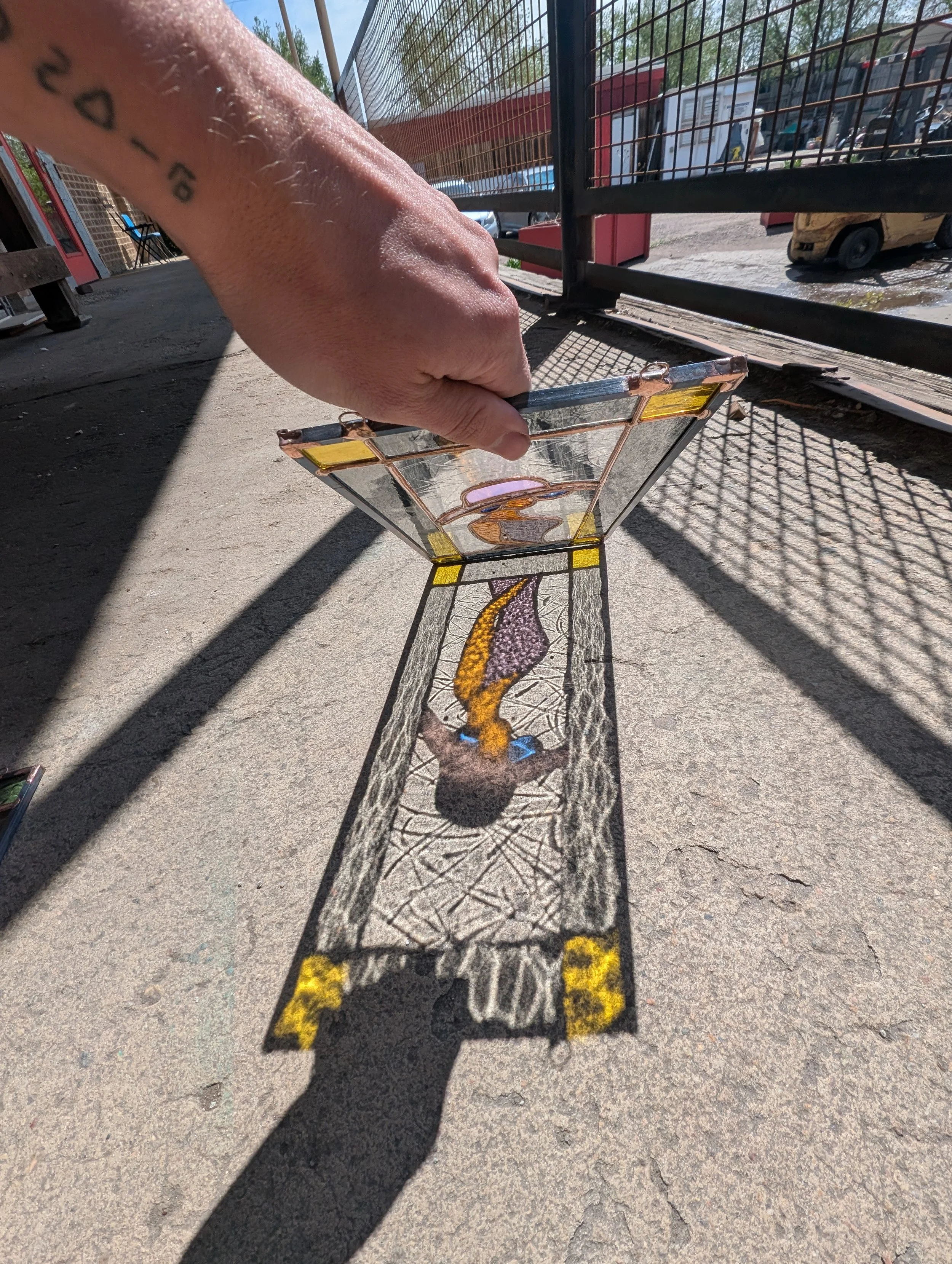 A person holding a stained glass panel with their right hand, casting a shadow that aligns with the artwork on the ground, depicting a nude woman with long hair, standing with her back to the viewer, in an outdoor setting.