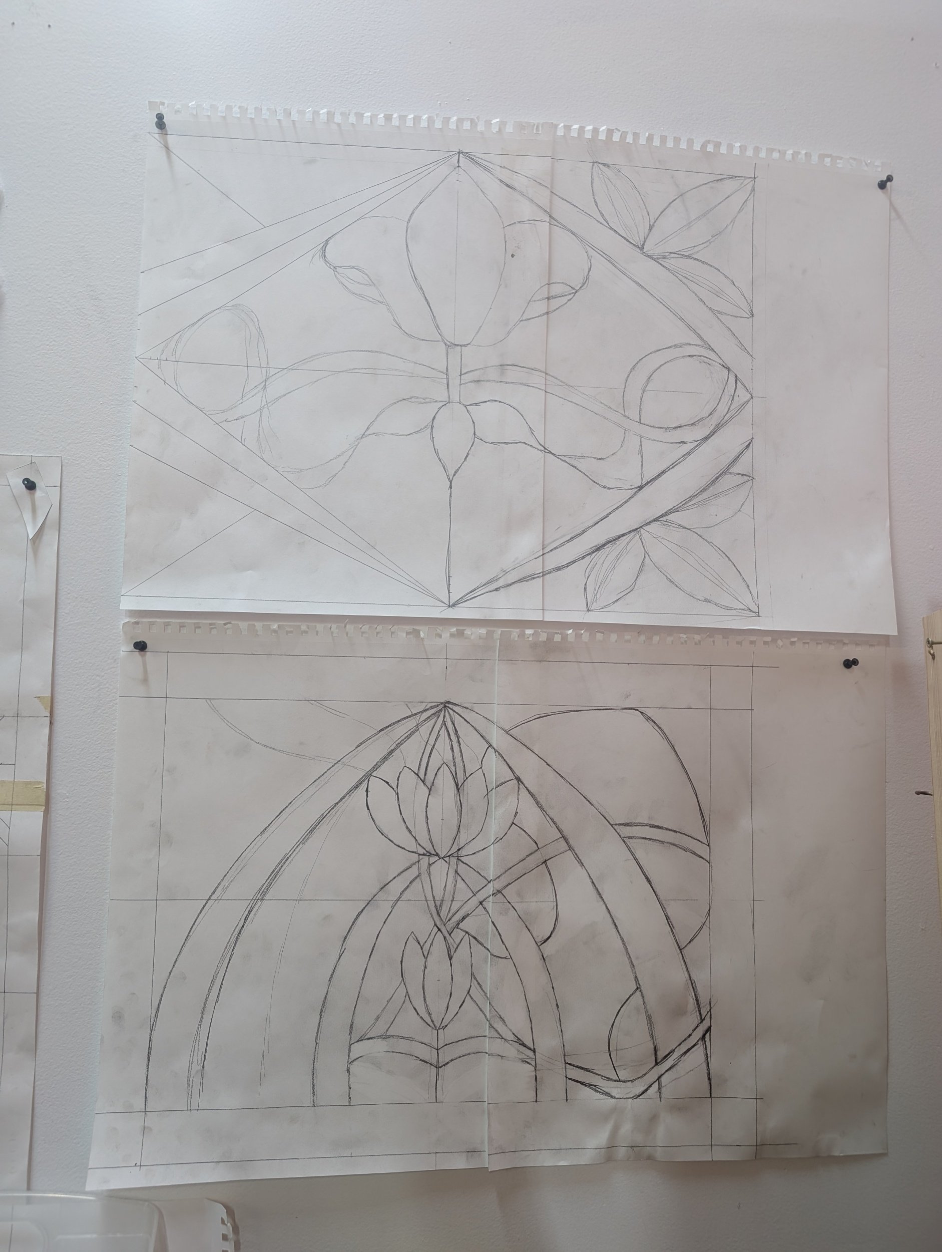 Two large sheets of paper with pencil sketches of stained glass window designs pinned on a wall, featuring floral and geometric patterns.