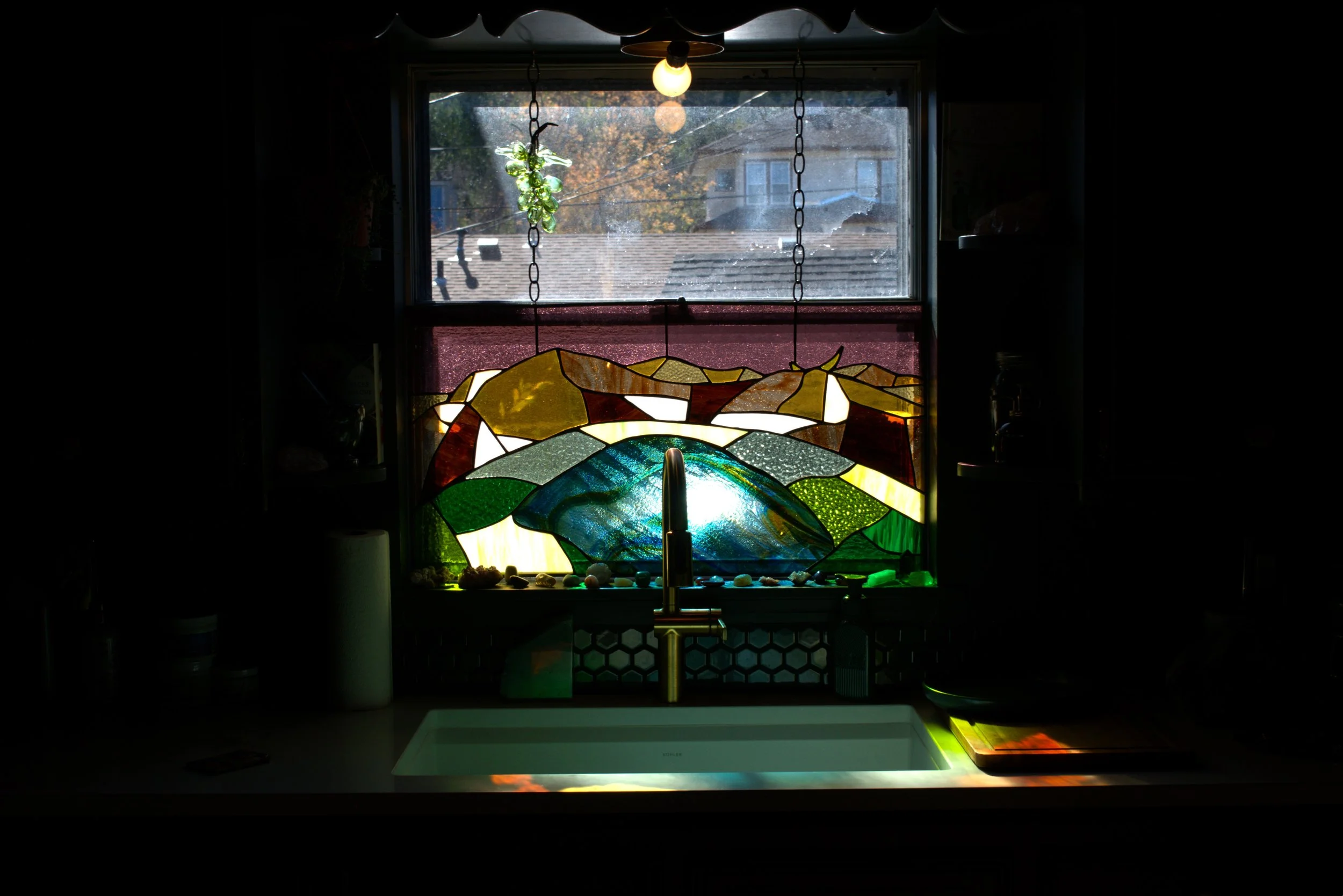 A kitchen window with stained glass depicting colorful mountains and sky, sunlight shining through, with a sink and faucet below.