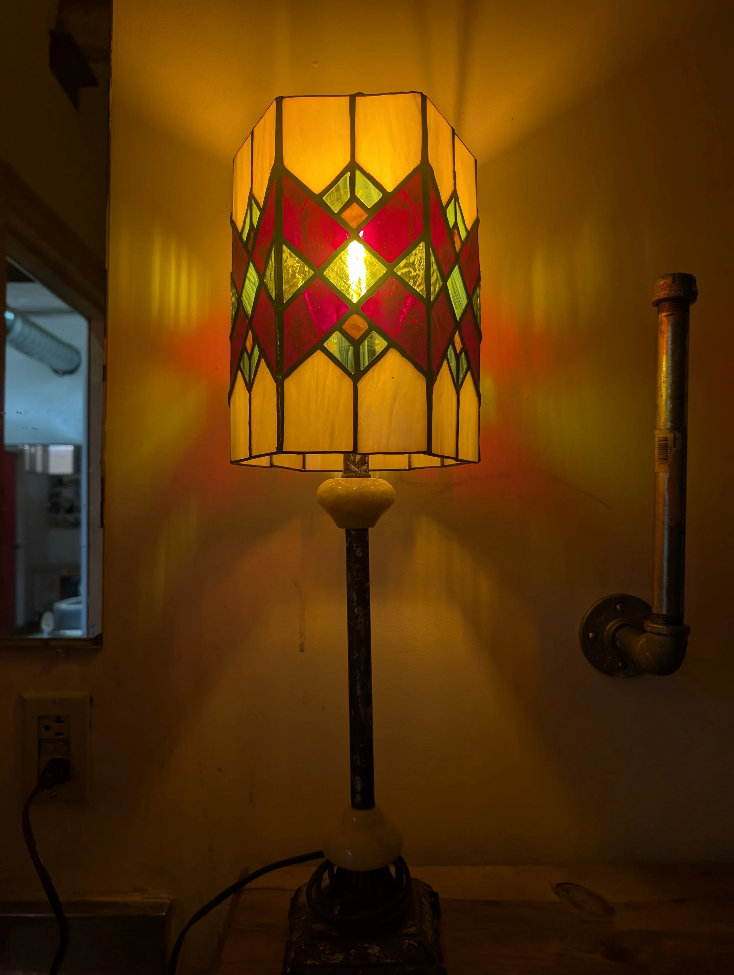 A stained glass table lamp with yellow, red, and green geometric patterns, lit and casting warm light on the wall behind it.