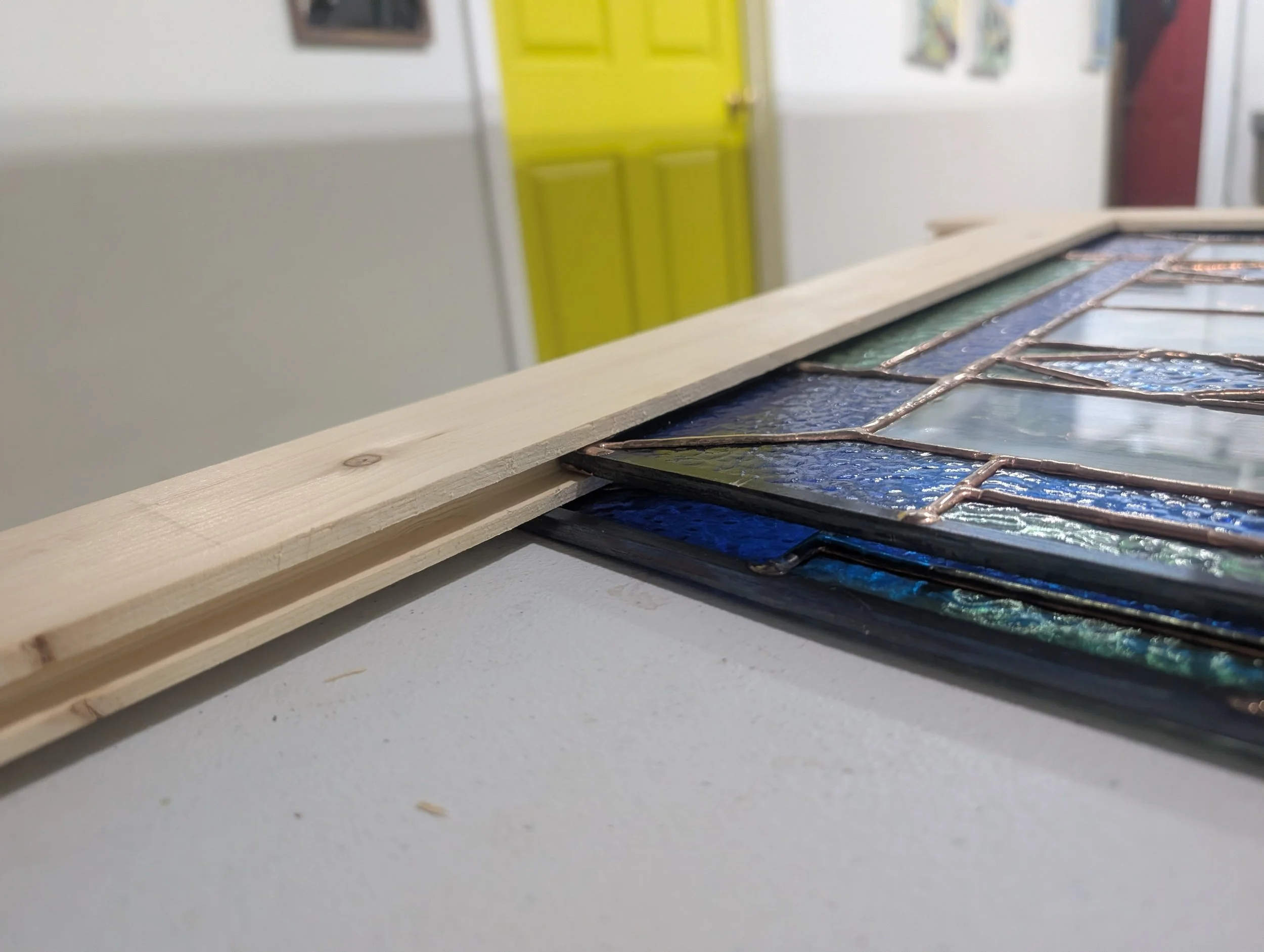 Close-up of a stained glass window piece on a white surface with a wooden strip on top, background shows a yellow door and some wall decorations.