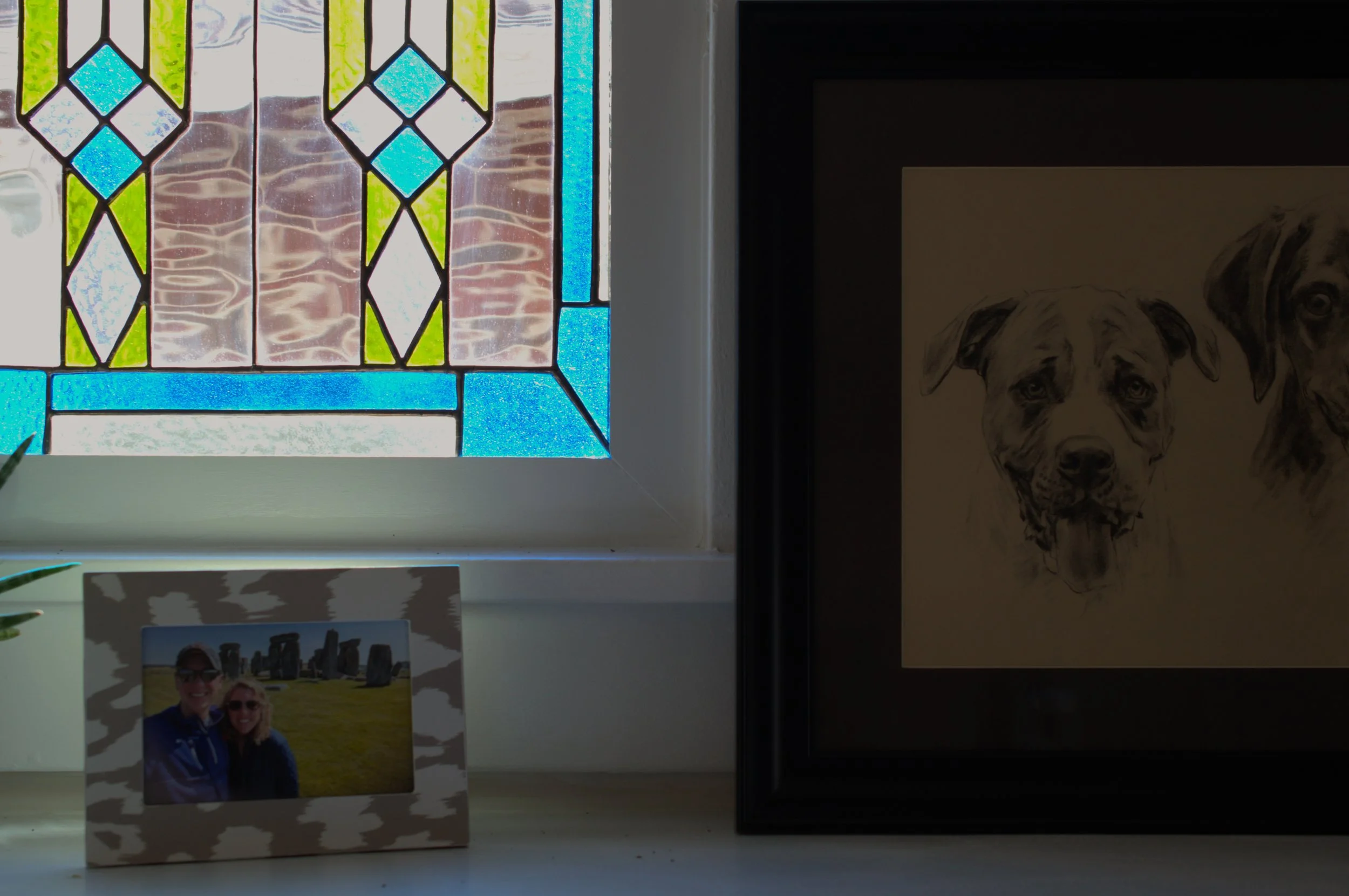 A framed photograph of a smiling couple standing outside with Stonehenge in the background, a black and white dog portrait, and a stained glass window with colorful geometric patterns on a windowsill.
