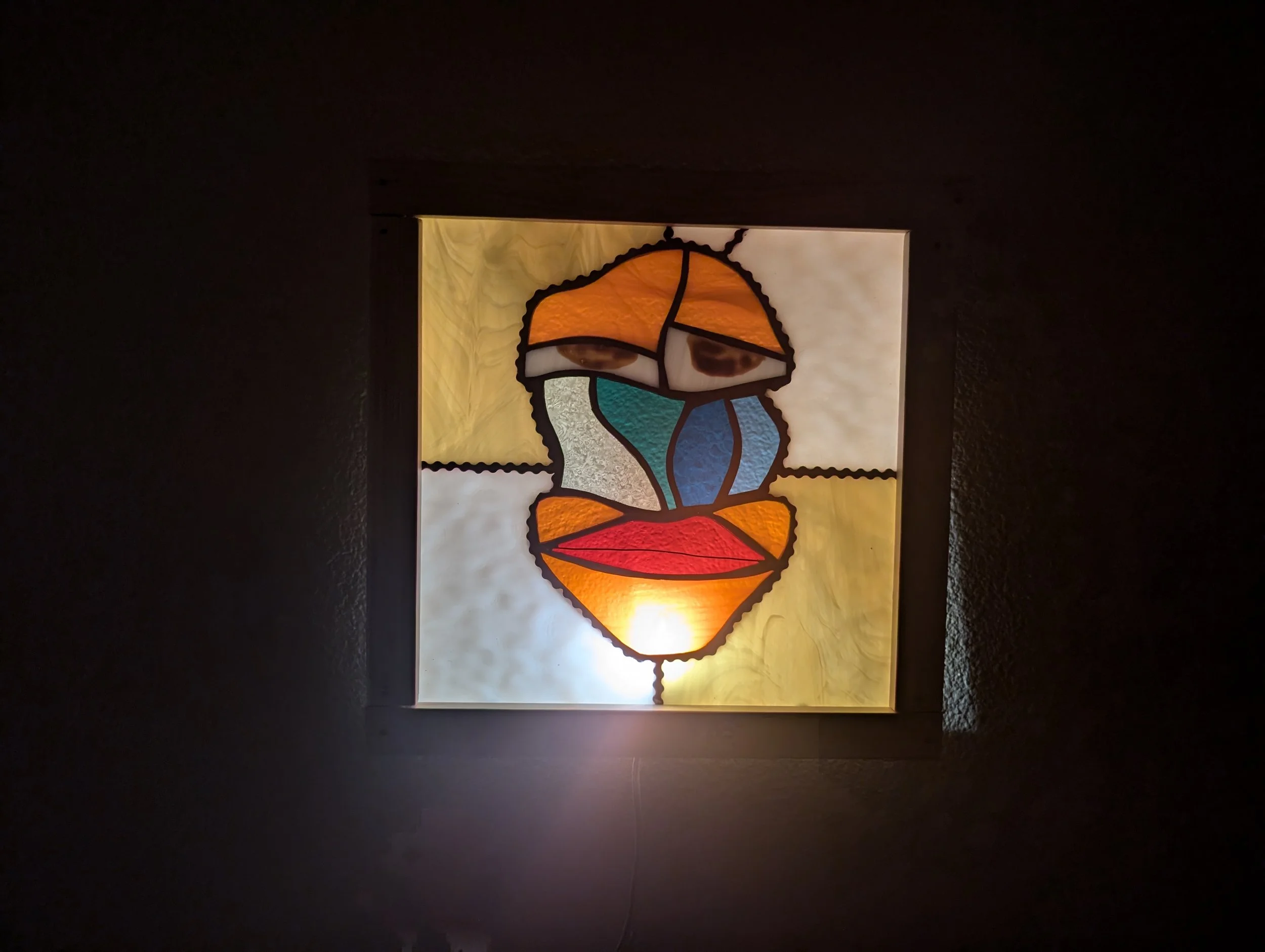 Colorful stained glass artwork featuring an abstract face with orange, blue, yellow, and red shapes, illuminated from behind.