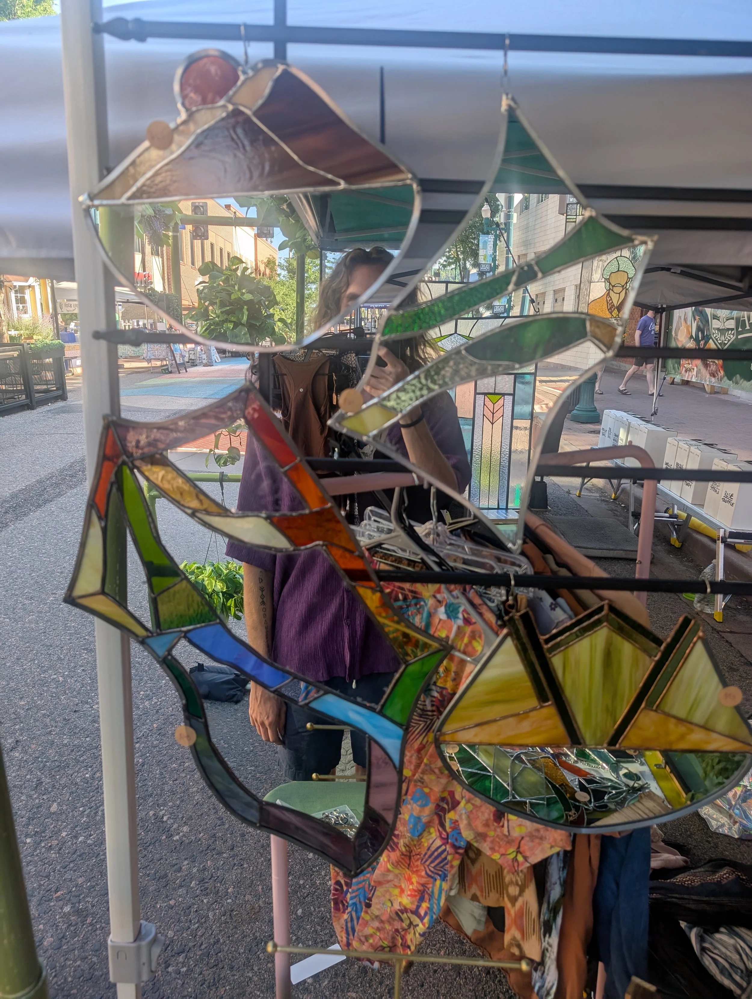 Colorful stained glass mirrors and decorations displayed at an outdoor market booth, with a reflection of a person taking a photo.