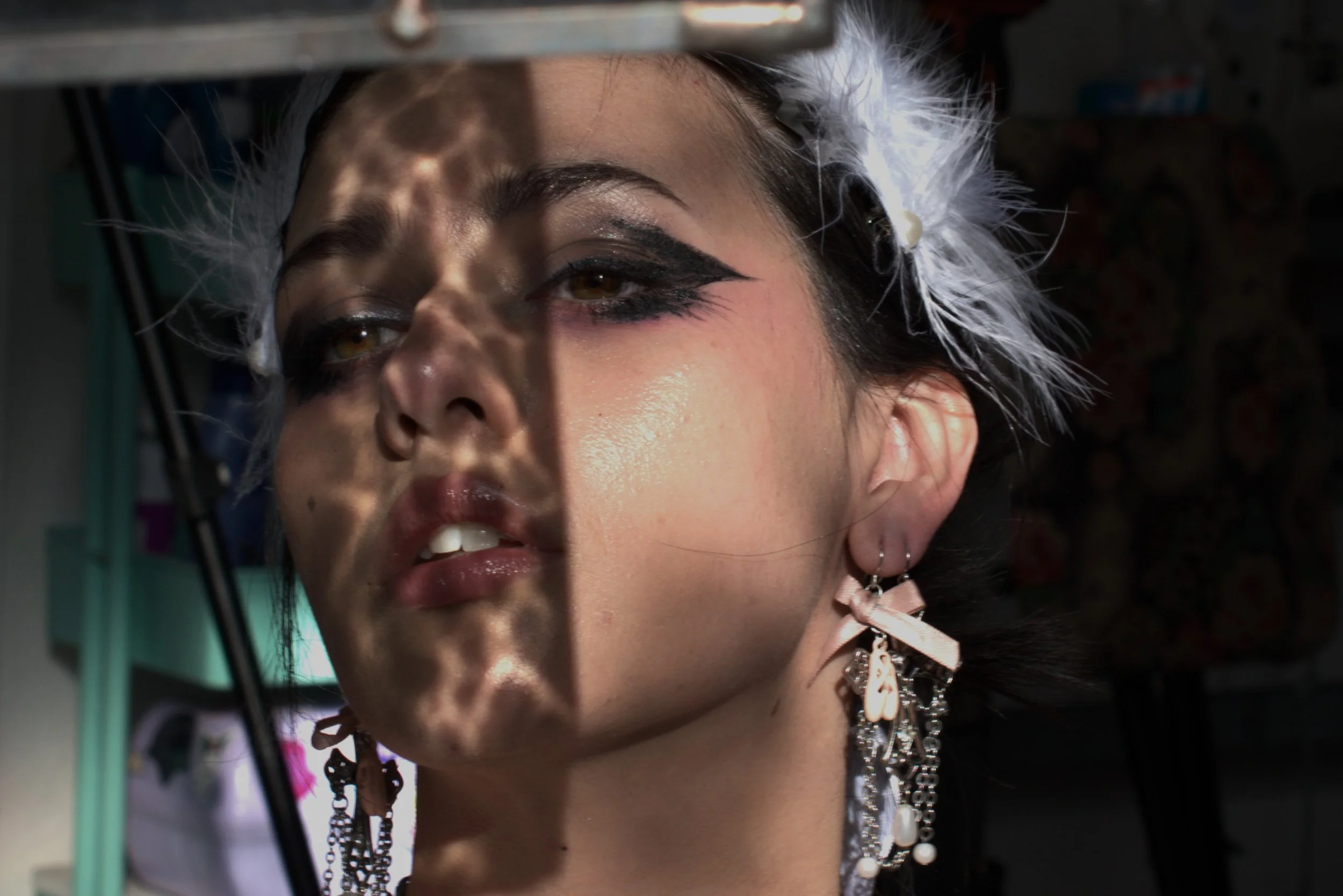 A woman with heavy eye makeup and earrings looks into a mirror, which reflects part of her face and creates a shadow pattern across her face.
