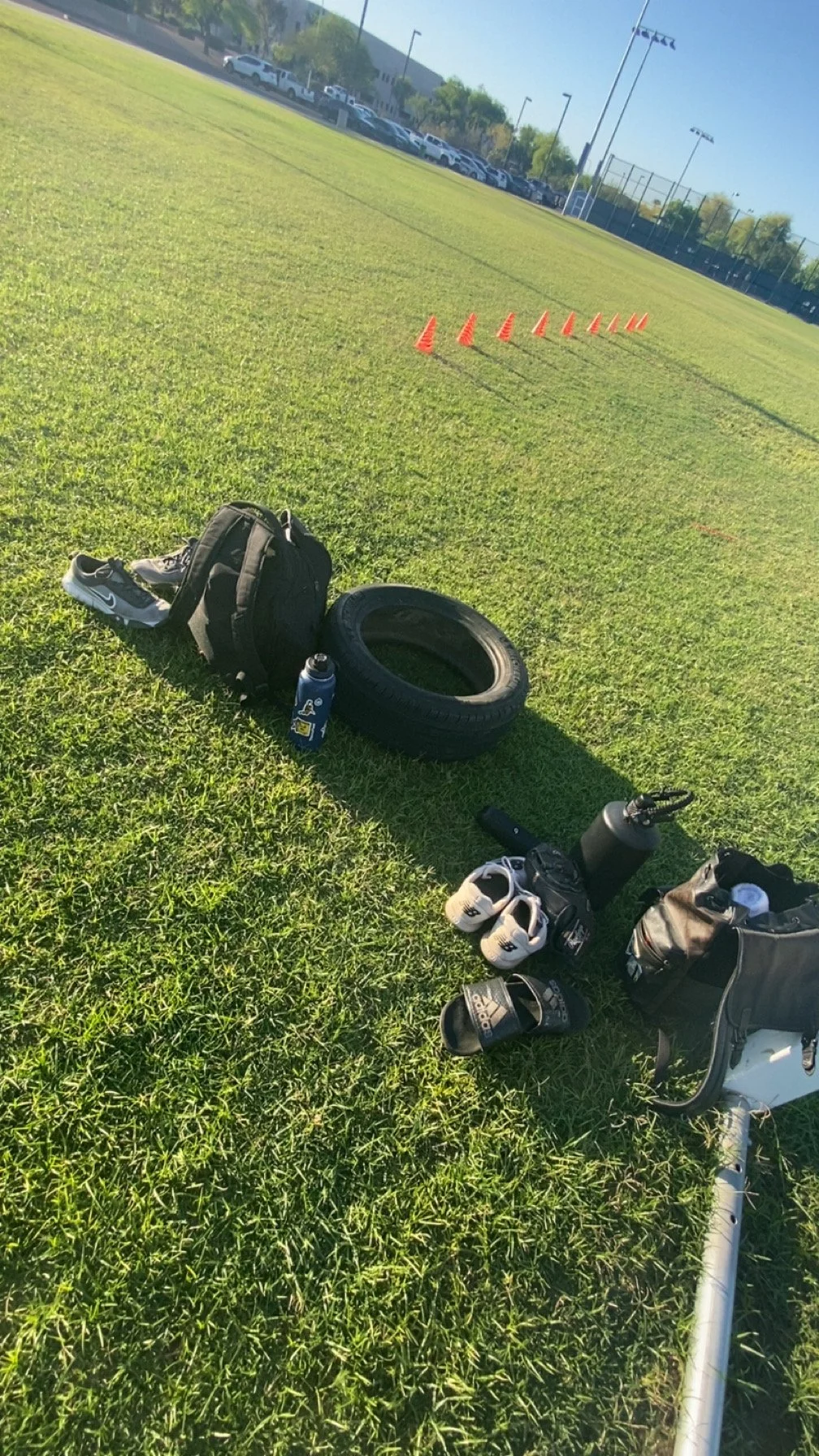 A sports field with tennis court fences and lighting poles in the background. Placement of orange cones on the grass, and a collection of athletic gear including shoes, water bottles, and a scooter on the ground.