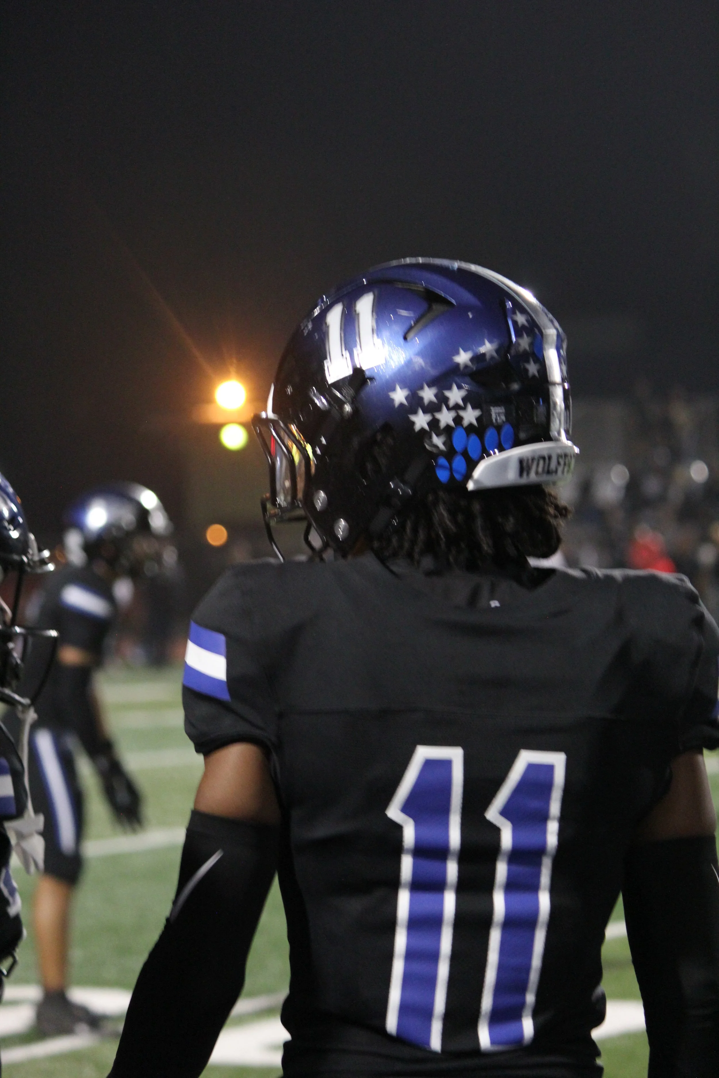 A football player wearing a black jersey with the number 11 and a blue helmet with stars, standing on the field at night.