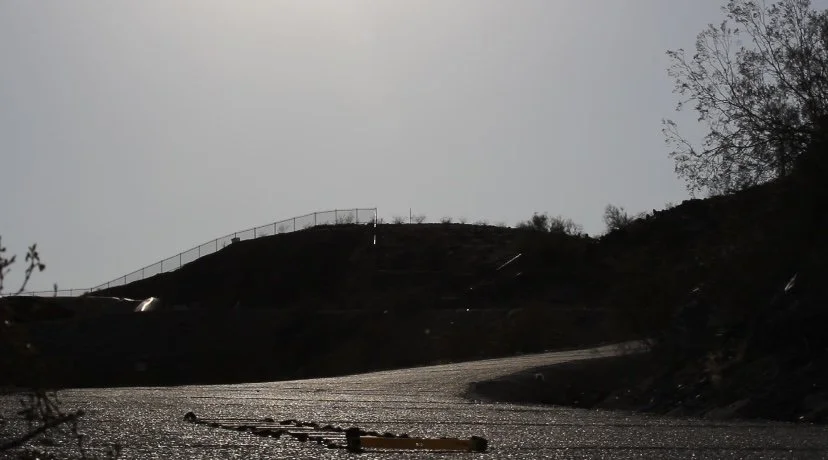 A winding road on a hilly terrain with trees and a fence on the top of the hill, under a cloudy sky.