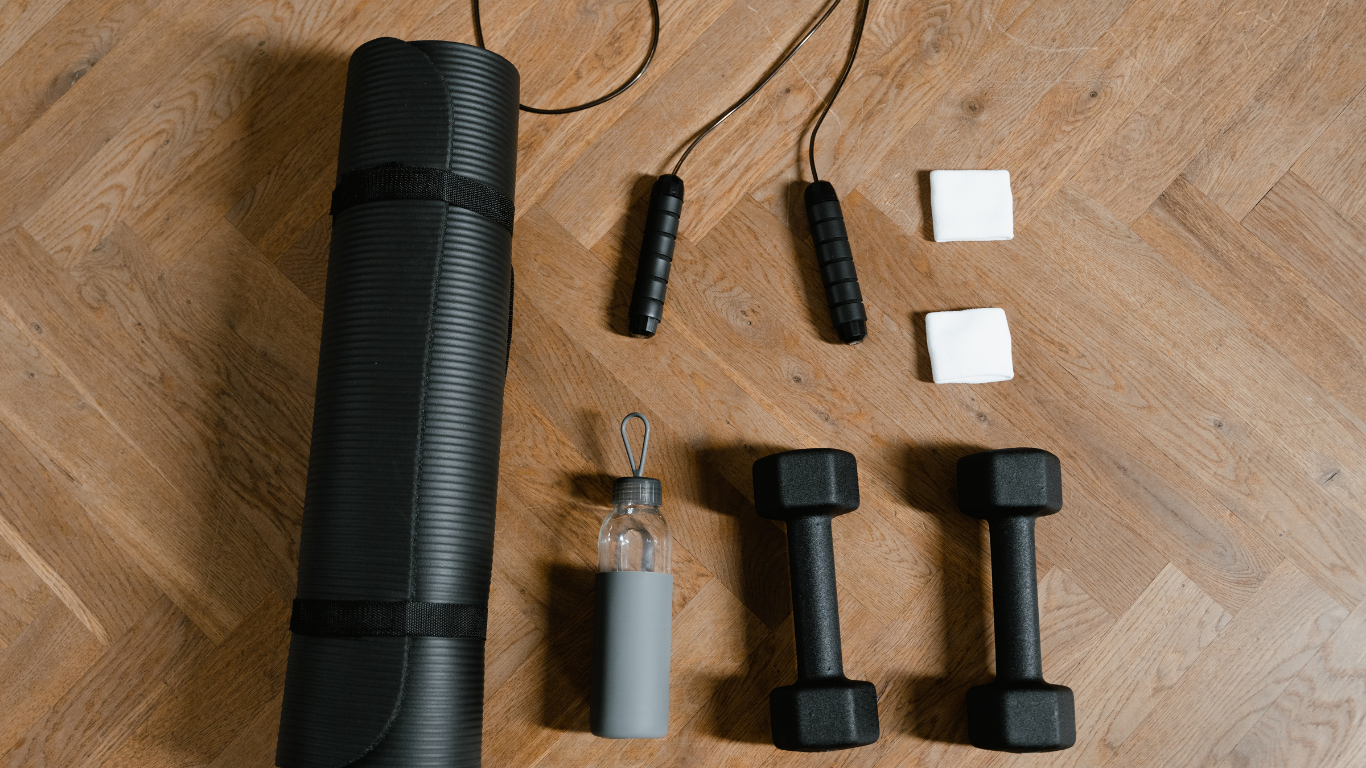 Home workout equipment on a wooden floor, including a black yoga mat in a rolled-up position, two black jump ropes, two white foam blocks, a gray water bottle with a loop top, two black dumbbells, and a pair of small white blocks.