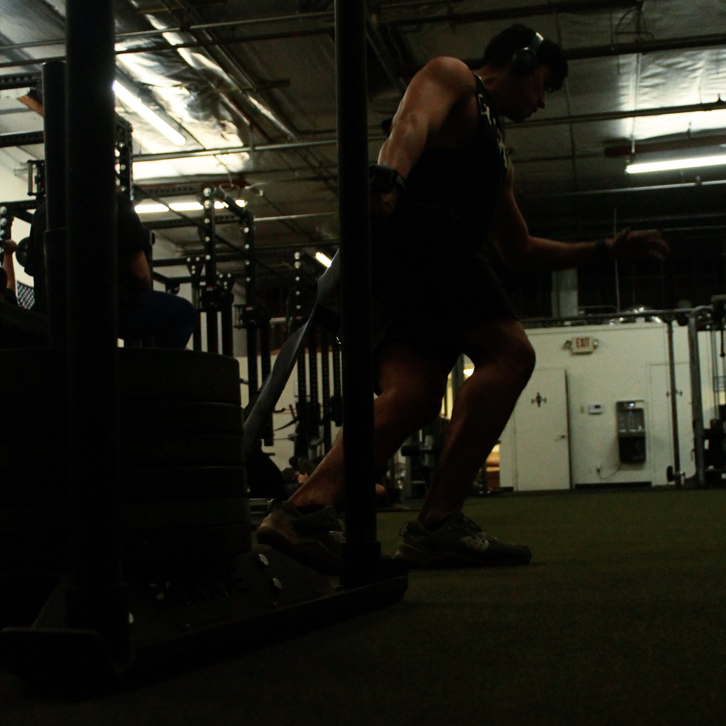 A man wearing headphones and workout attire performing a squat exercise with a weight attached to his waist in a gym.