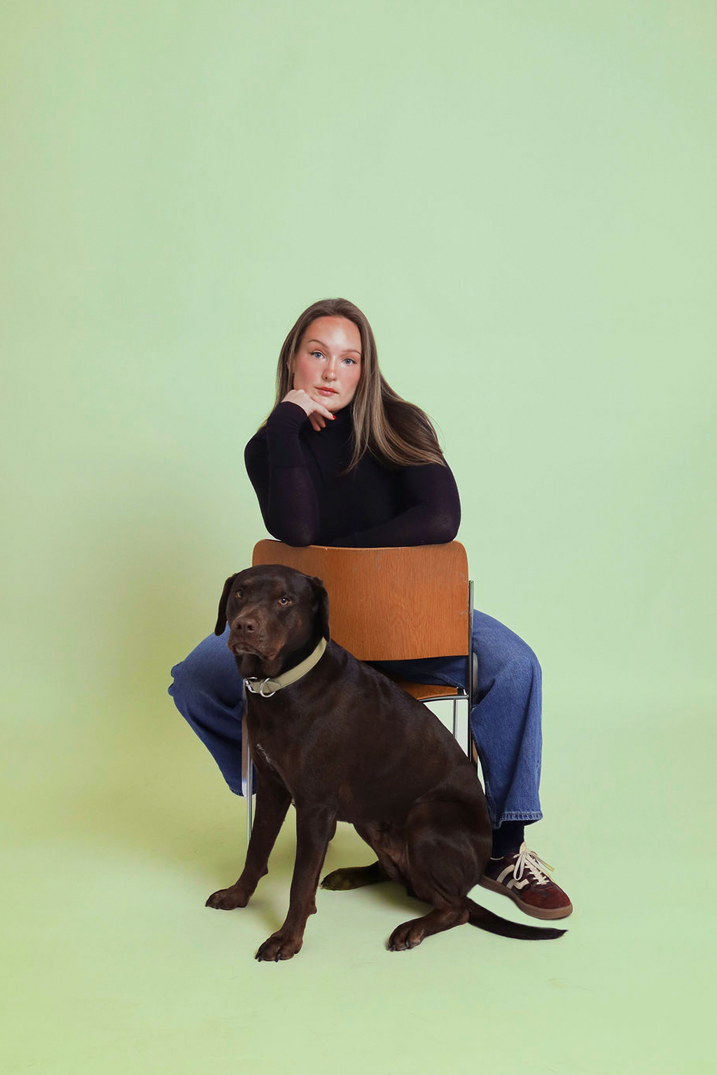 A young woman with long brown hair sitting on a wooden chair with a brown Labrador retriever in front of her, against a pale green background.