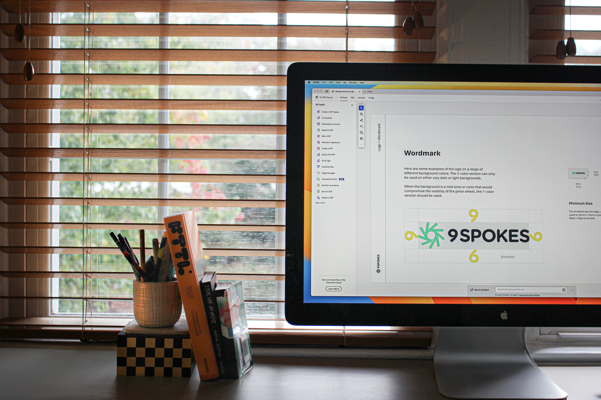 A computer monitor displaying a design guide for a logo called '9Spokes' with a green abstract spiral symbol and colorful numbers, placed on a white desk with some books, pens, and a window with wooden blinds in the background.