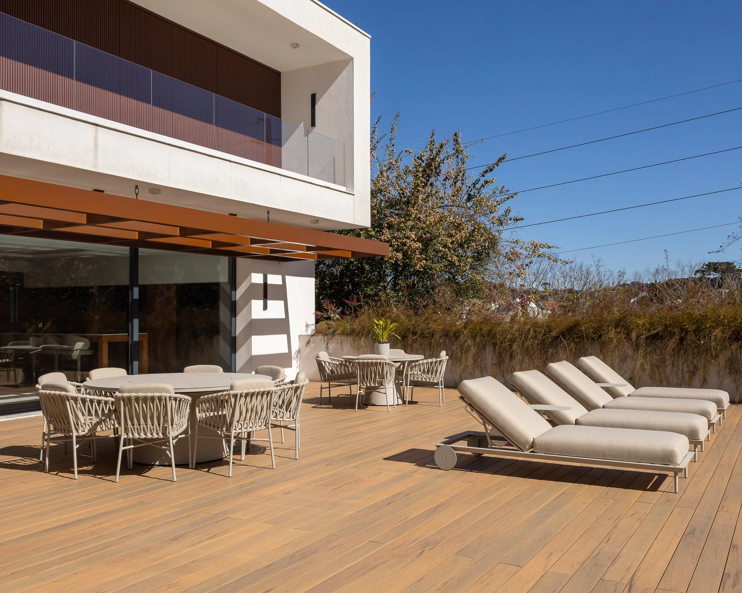 Deck de madeira com espreguiçadeiras, mesa de jantar com cadeiras ao ar livre, e uma planta ao centro, com uma casa moderna ao fundo e céu claro.