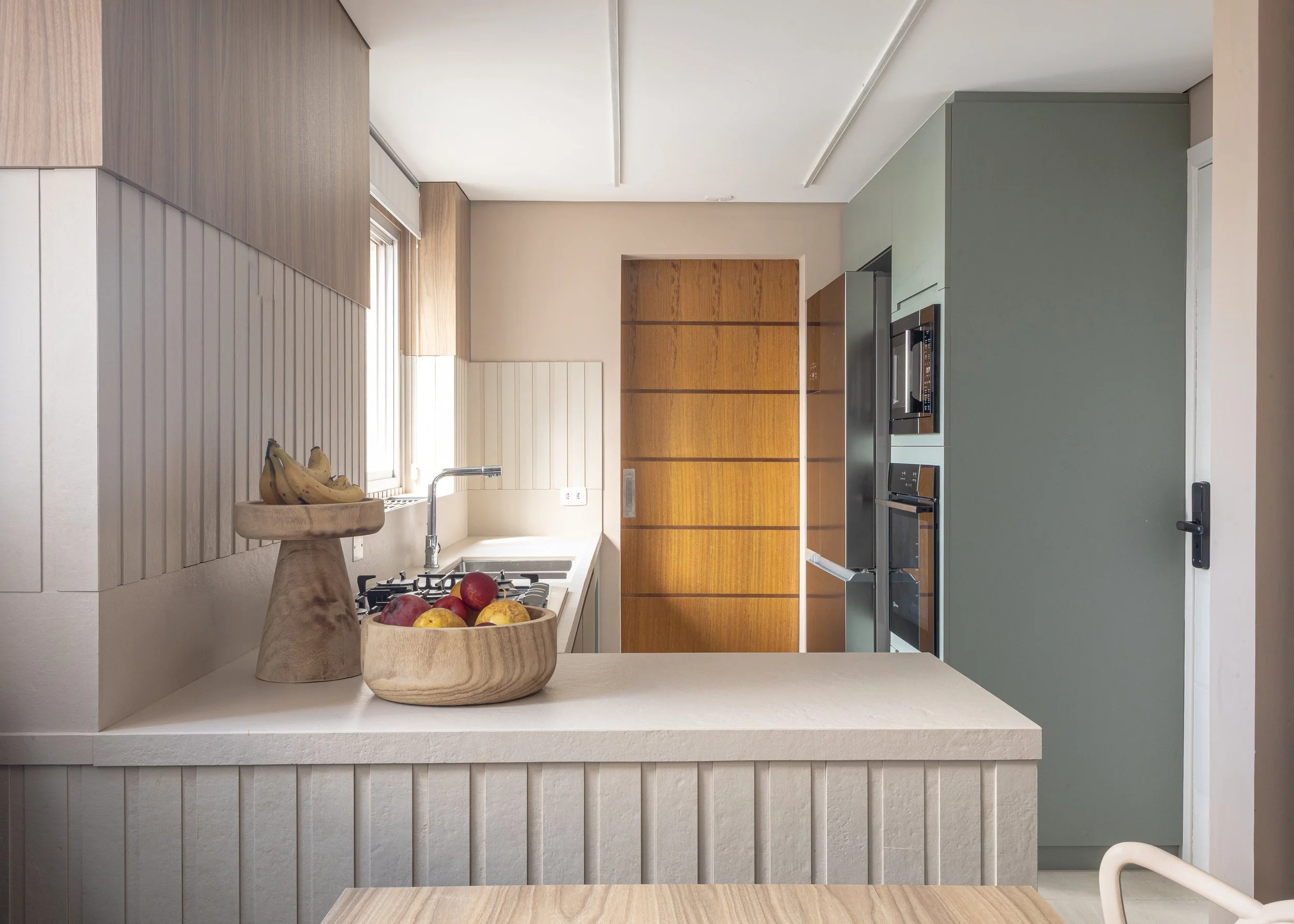 Cozinha moderna com armários de madeira, bancada de granito, frutas em uma tigela de madeira e janela com cortinas brancas.