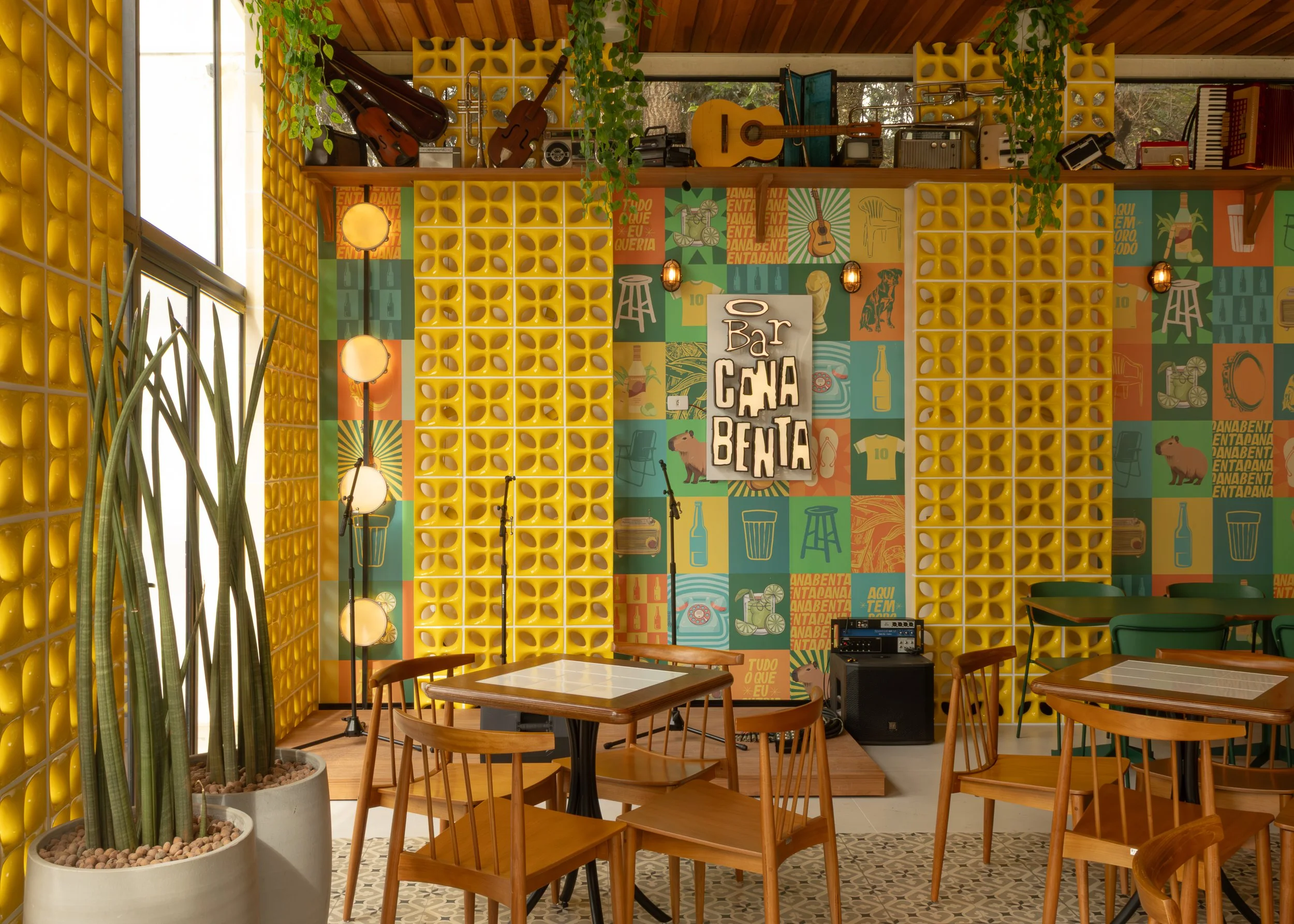 Interior de bar com decoração colorida, paredes decoradas com quadrados amarelos de efeito 3D, poster com o nome 'Bar Canoa Bentana', instrumentos musicais no fundo, como violões e violino, plantas penduradas, mesas de madeira com cadeiras de madeira, iluminação com lâmpadas de filamentro exposto, e piso de azulejos decorativos.