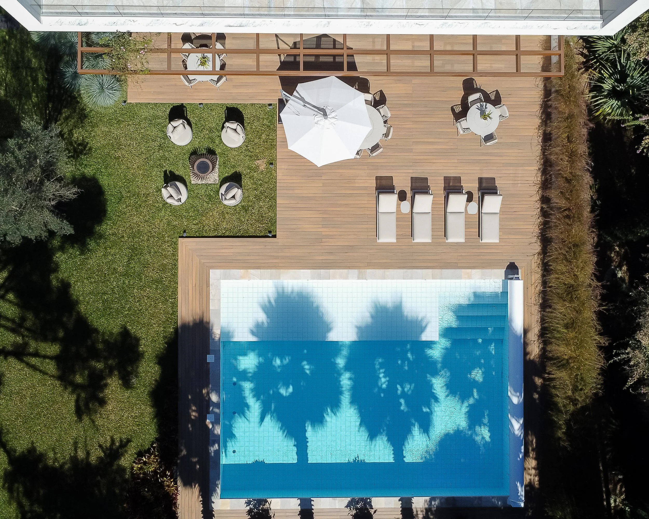 Vista aérea de uma área de lazer com piscina, espreguiçadeiras, mesas com guarda-sol, cadeiras em uma varanda de madeira e área de jardim com grama e árvores.