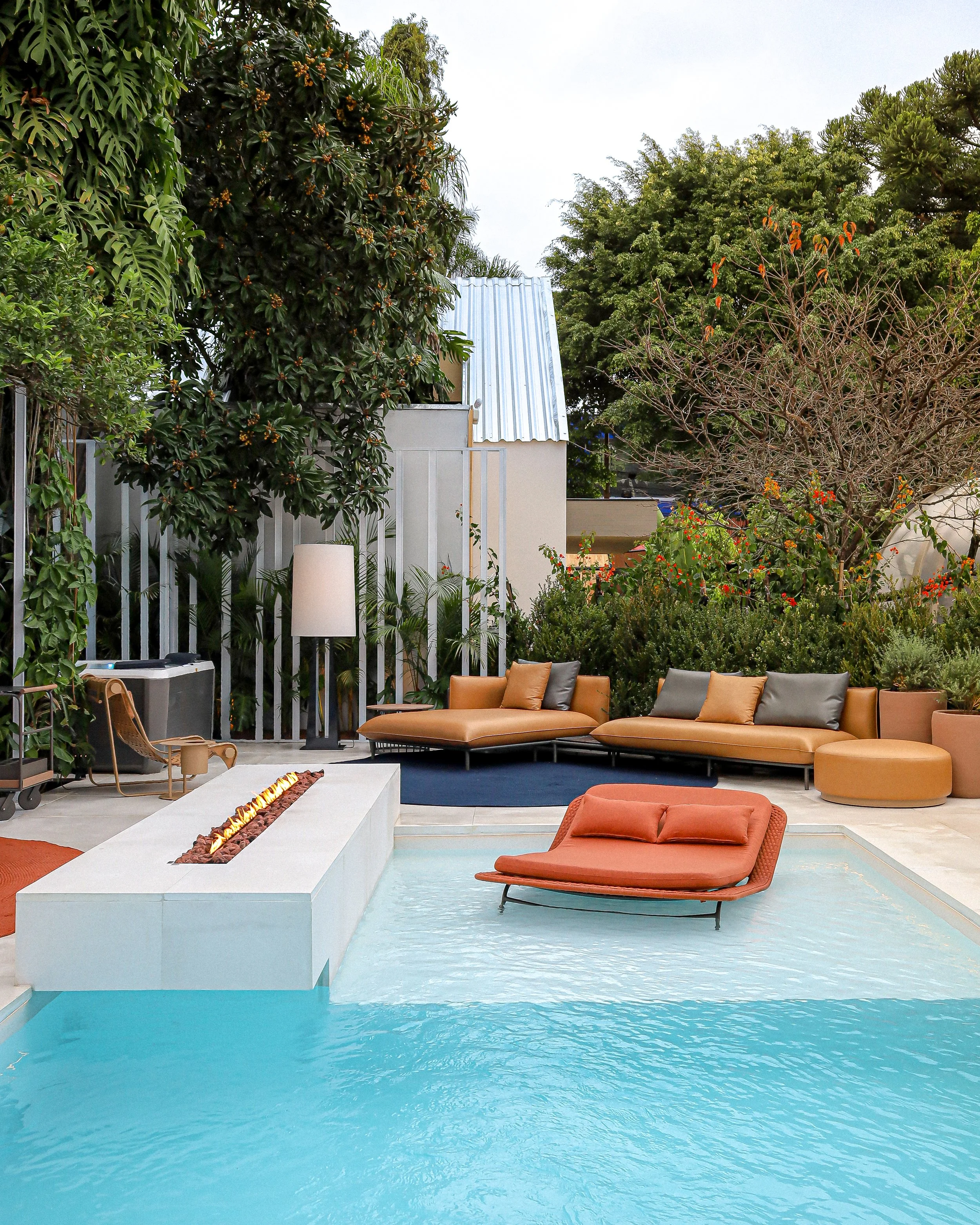 Área de piscina com espreguiçadeiras laranjas, uma delas parcialmente submersa na água, ao lado de uma lareira de fogo na borda da piscina, mobiliário de exterior confortável rodeado por árvores e vegetação.