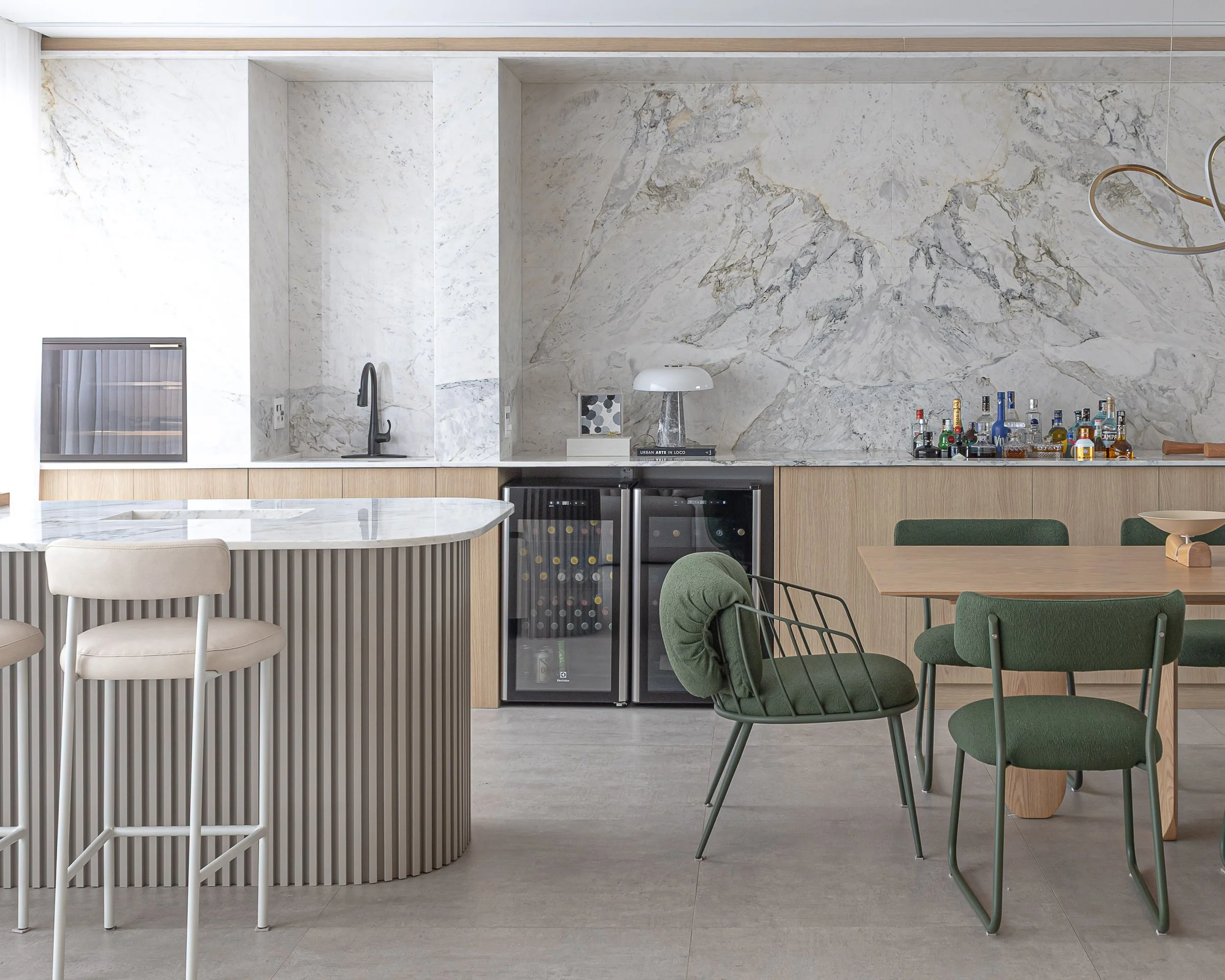 Cozinha moderna com parede de mármore, balcão de madeira, geladeiras de vinho, bancada com pia, iluminação discreta, mesa de jantar com cadeiras verdes, decoração minimalista.