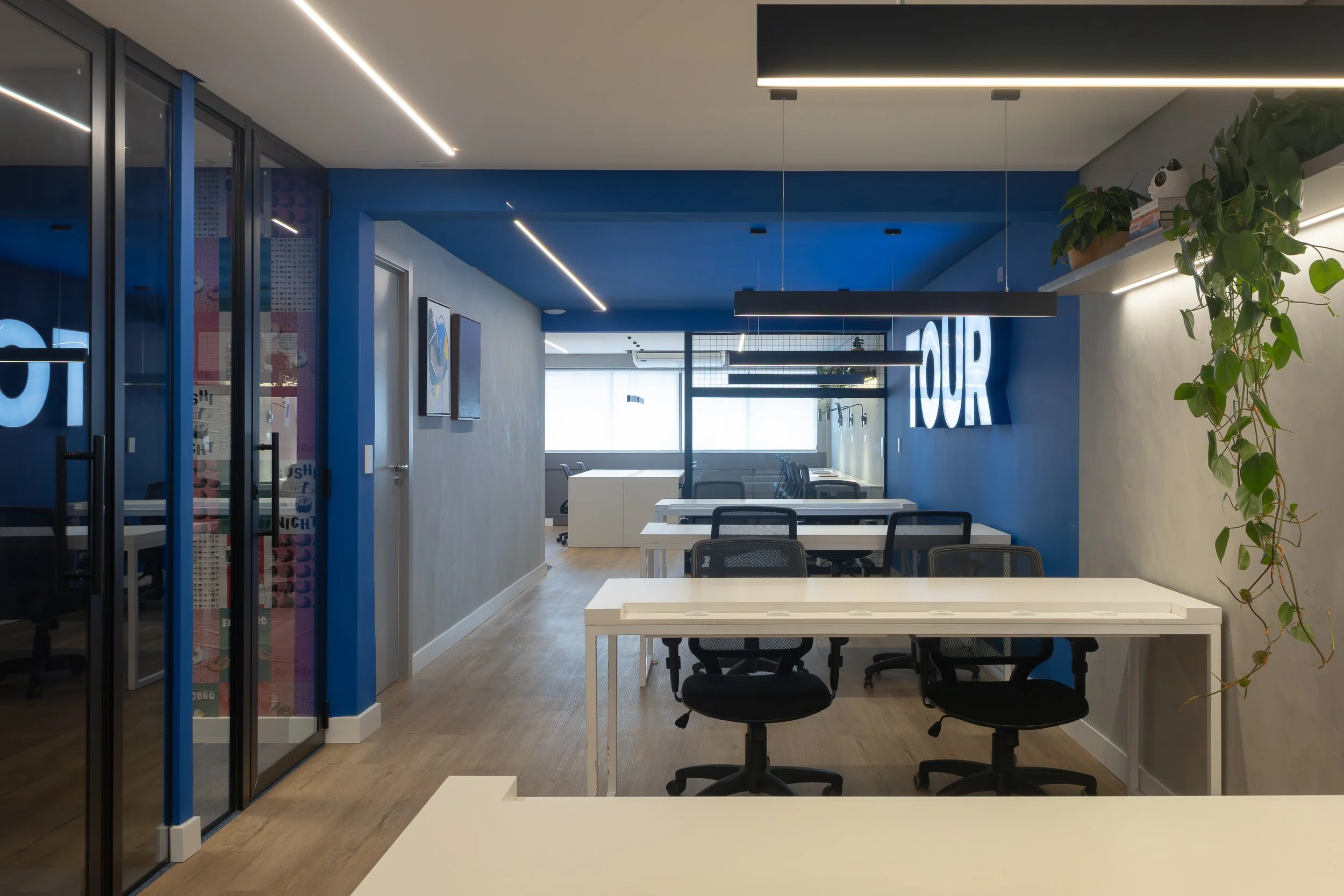 Escritório moderno com mesas de trabalho, cadeiras pretas, paredes cinza, uma parede azul com a palavra 'TOUR' iluminada, plantas e iluminação linear no teto.