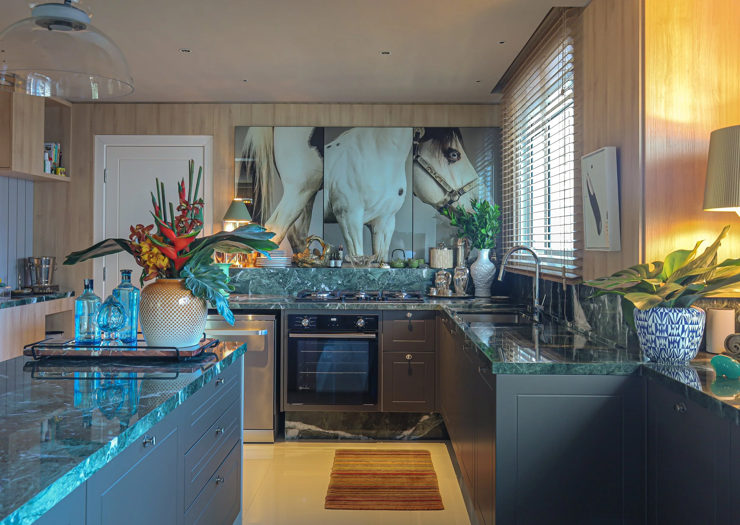 Cozinha moderna com bancadas de mármore verde escuro, armários cinza, janela com persianas, decoração com vasos de plantas, quadro grande de cavalos na parede e utensílios de cozinha.