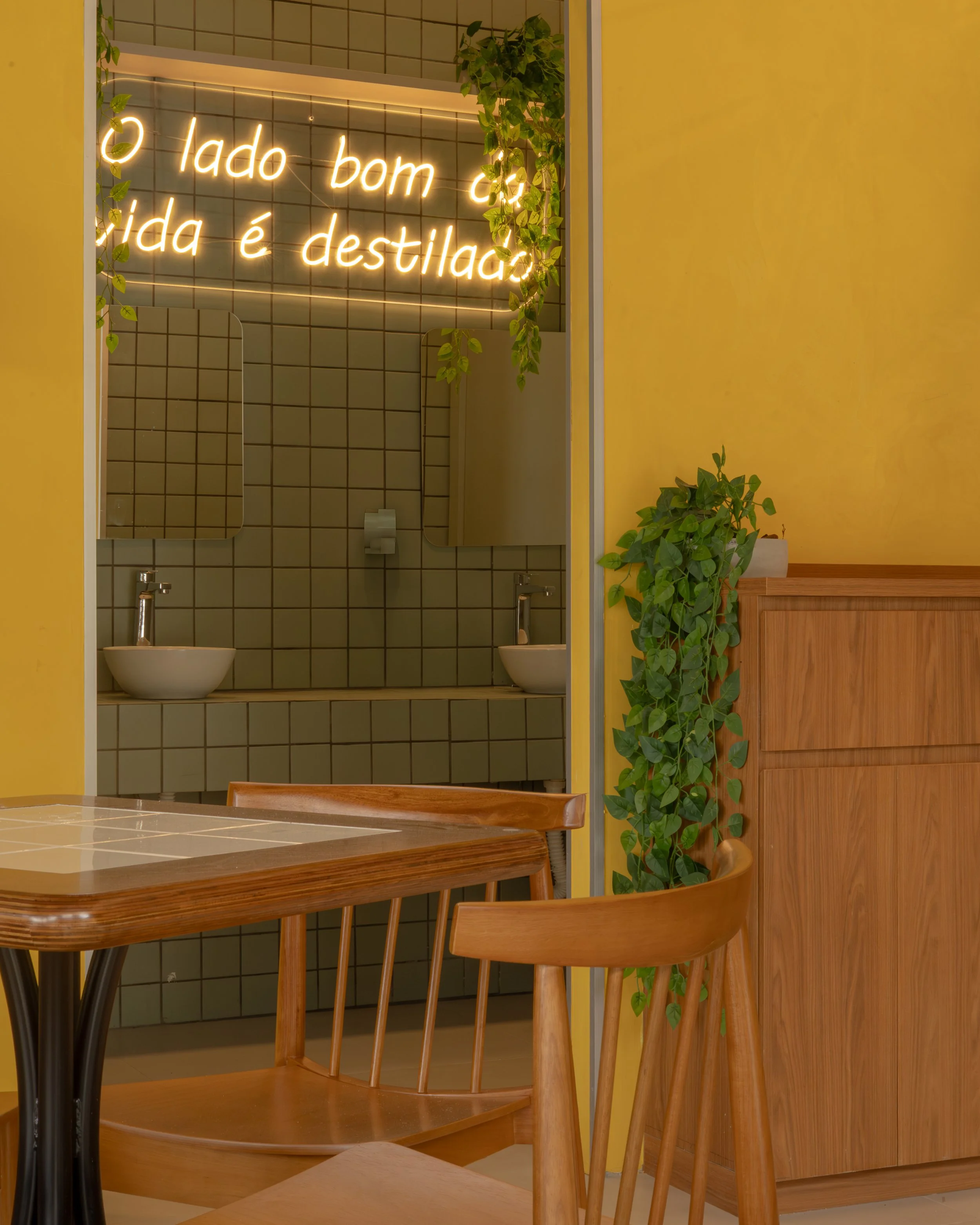 Interior de um restaurante com parede amarelo, uma mesa de madeira com cadeiras também de madeira, uma divisória com plantas e um espelho, e uma parede de azulejos com uma luminária de neon que exibe uma frase em português.