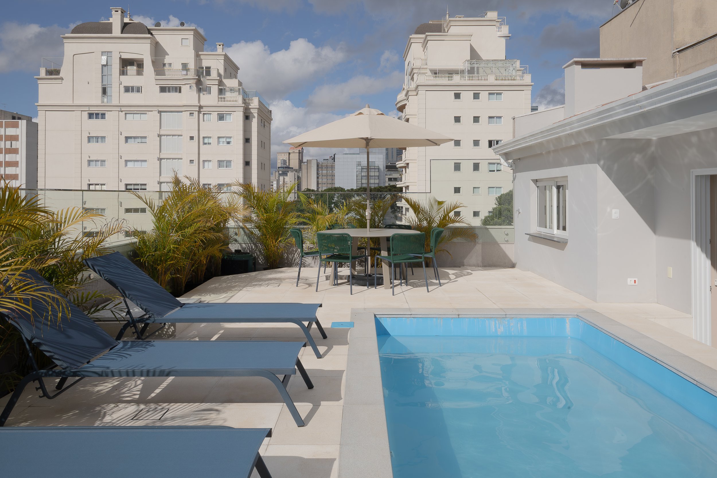 Vista de uma piscina em um terraço com espreguiçadeiras azuis, uma mesa com guarda-sol e cadeiras verdes, plantas ao redor e prédios residenciais ao fundo, em um dia ensolarado com céu parcialmente nublado.