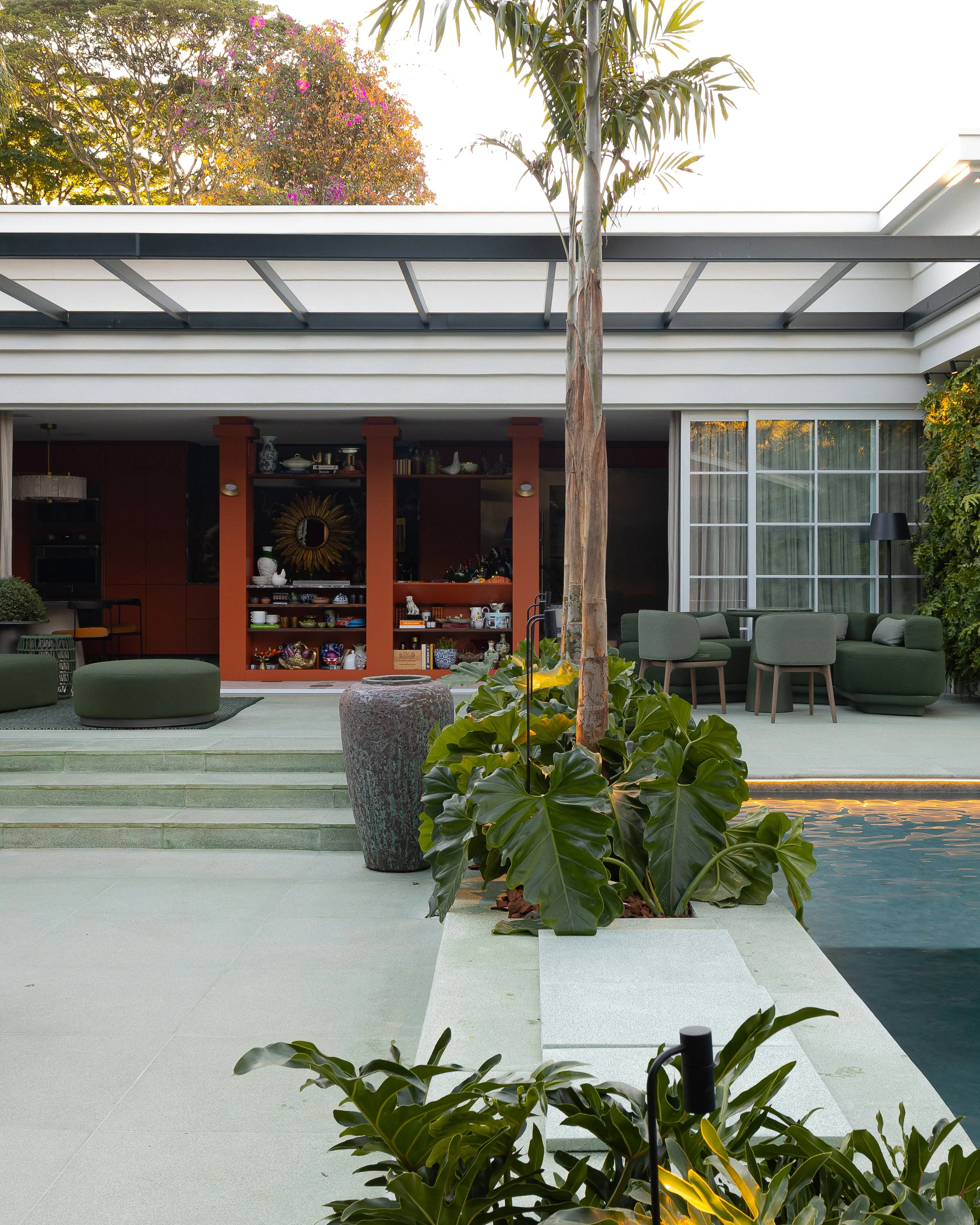 Imagem de uma área de lazer ao ar livre com piscina, árvores e plantas, incluindo uma palmeira, tendo uma varanda com uma área de estar com sofás e uma mesa, além de uma parede com prateleiras e uma porta de vidro.