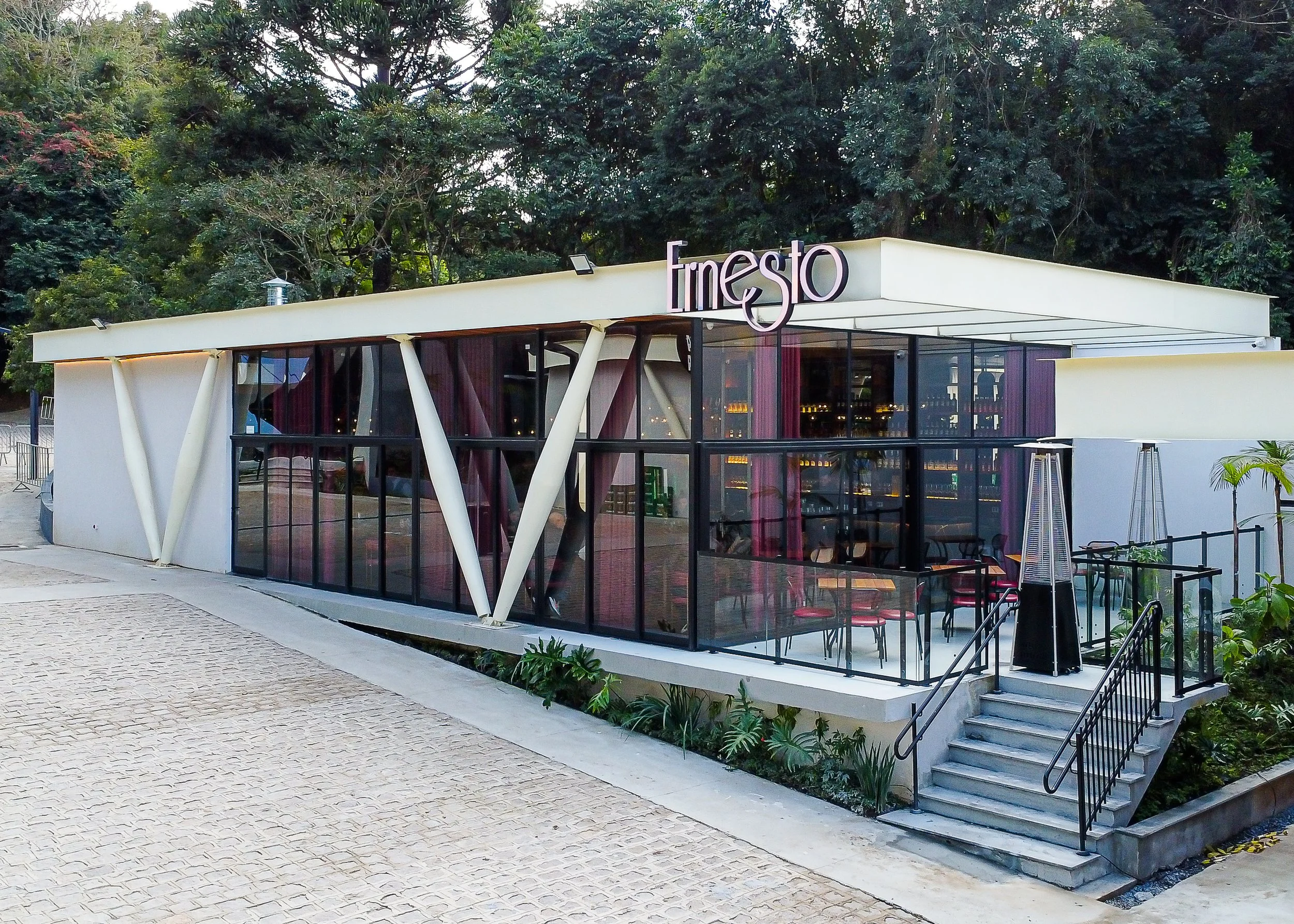 Fachada de um restaurante com nome 'Fresto', estrutura moderna com vidro e ilhas de vidro na varanda, cercado por vegetação e árvores ao fundo.