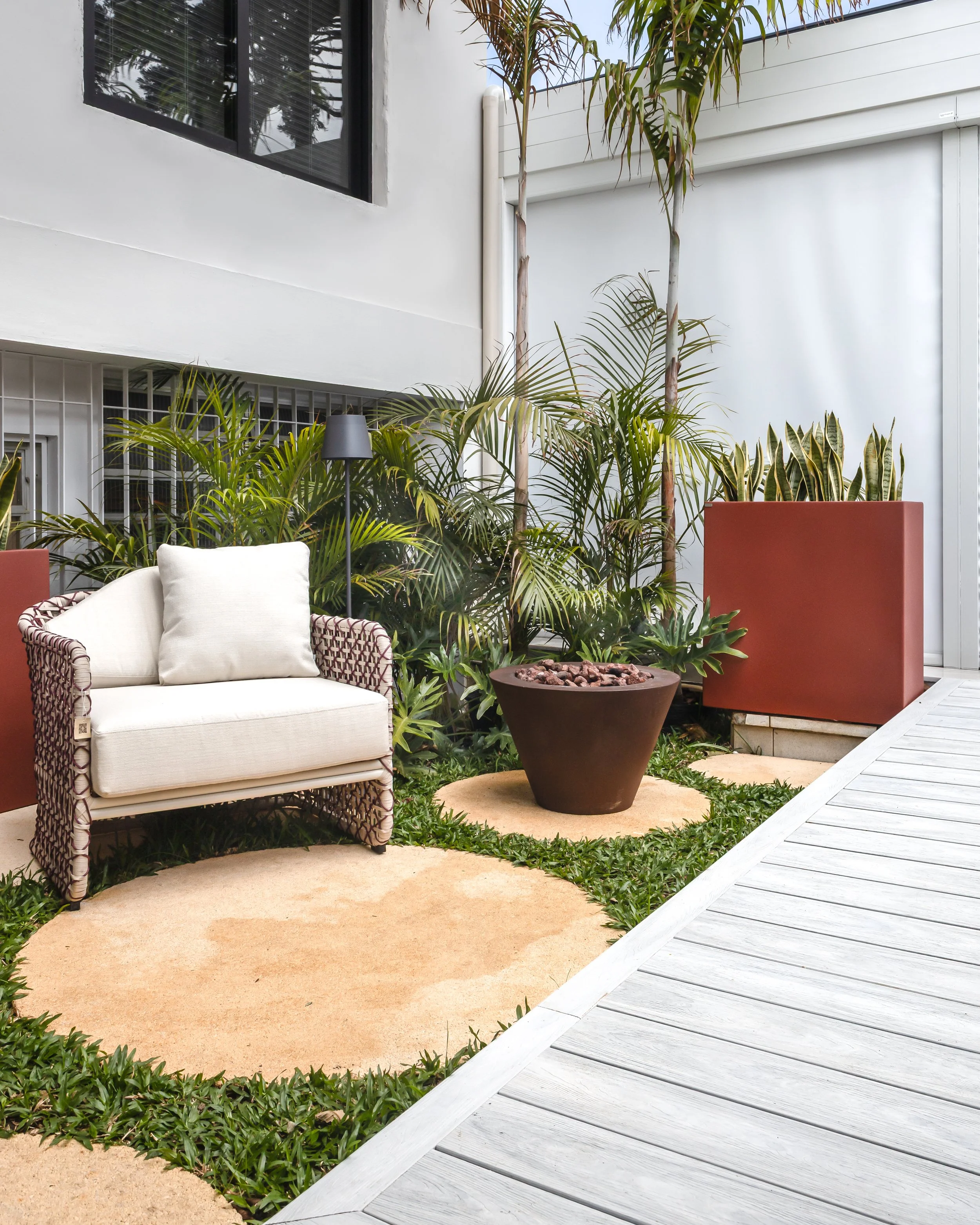 Espaço externo com área de descanso, contendo uma poltrona de tecido branca com estrutura decorativa de vime, uma luminária de pé preta, várias plantas verdes ao fundo, uma grande urna de barro com pedras, e uma parede branca com janela preta.