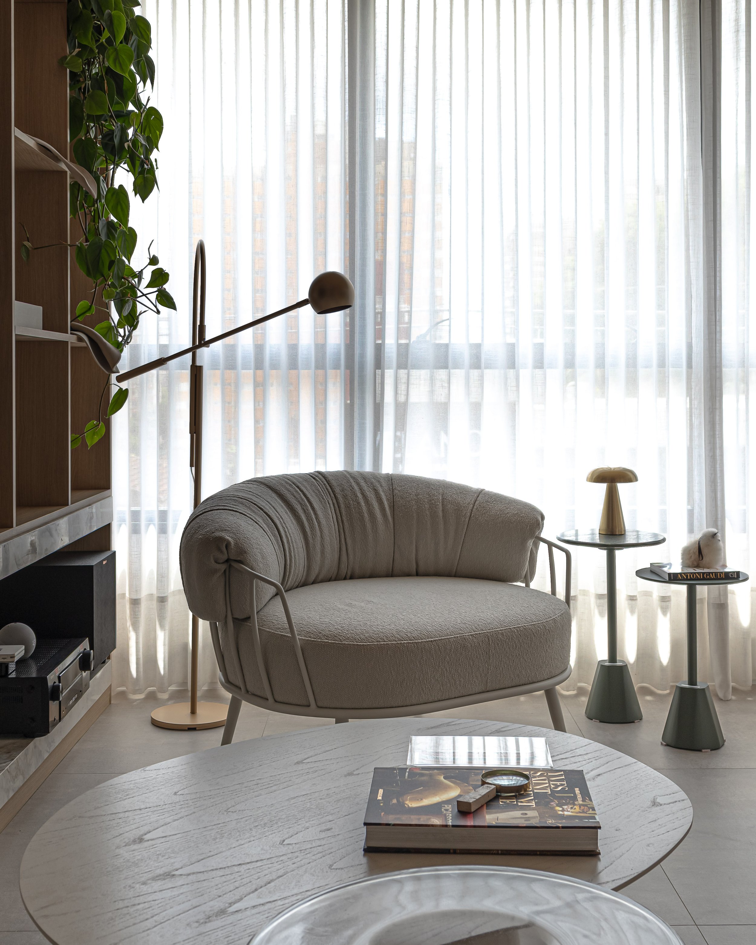 Sala de estar com sofá arredondado de tecido bege, mesa de centro de madeira clara, e janelas com cortinas brancas finas permitindo luz natural. Há luminária de chão ao lado do sofá, duas mesas laterais com itens decorativos e livros, e uma planta ao lado da janela.