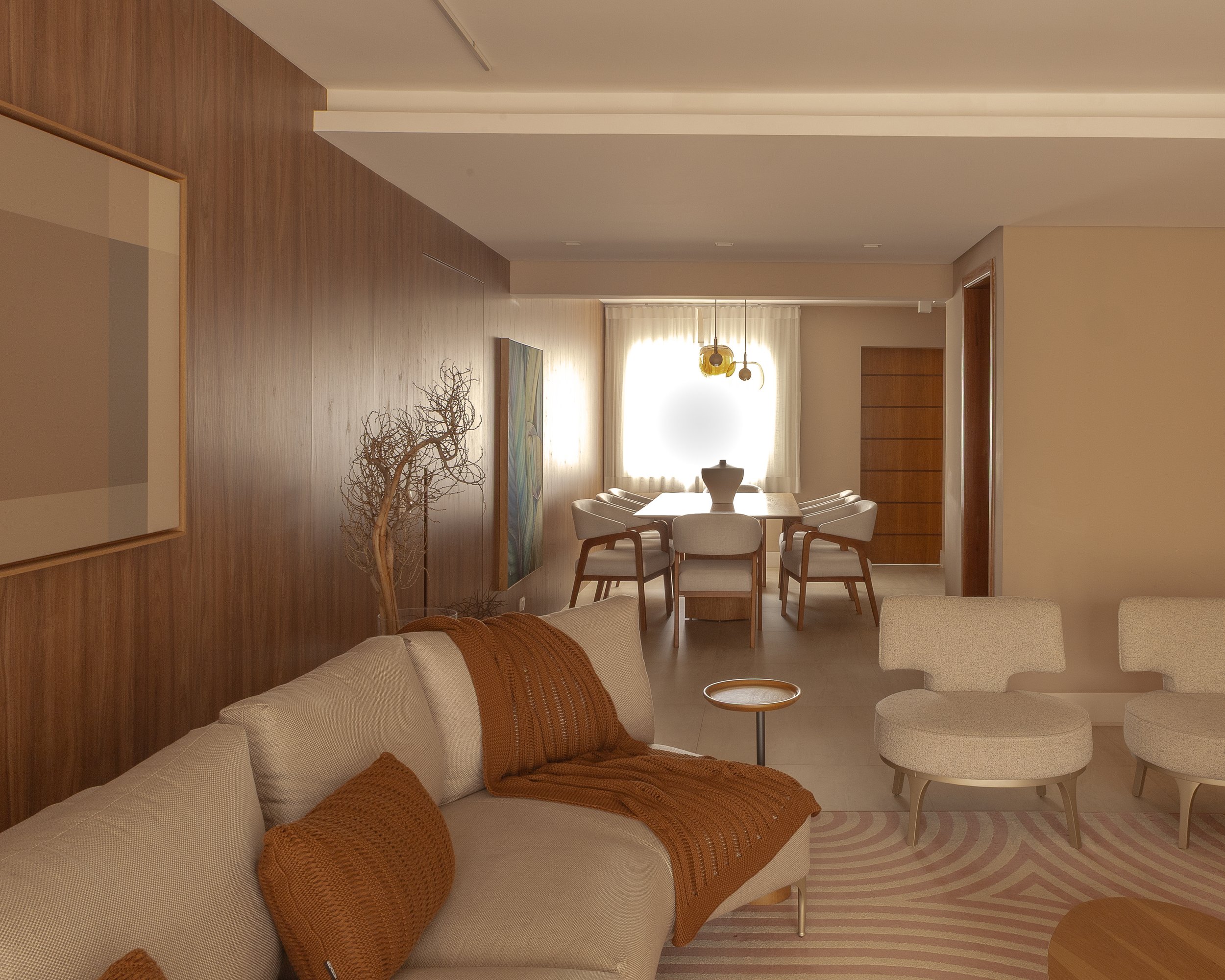 Sala de estar com sofá bege, cadeiras brancas, mesa de centro, decoração com elemento de tronco seco, pintura moderna na parede de madeira, iluminação natural das janelas, e uma área de jantar ao fundo com mesa e cadeiras