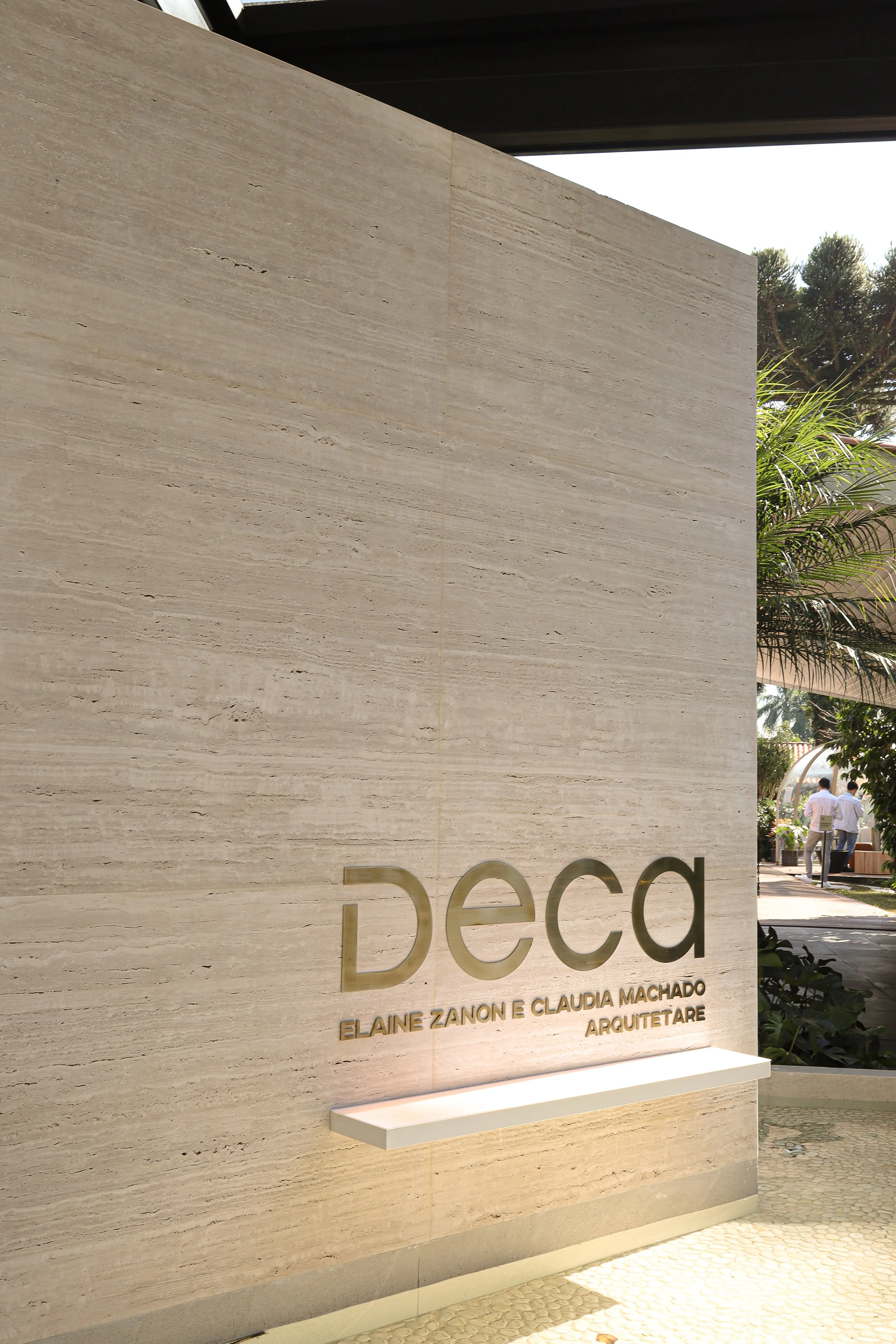 Placa de nome do escritório de arquitetura 'Deca' com os nomes Elaine Zanon e Claudia Machado gravados em relevo, fixada em parede de pedra claro na entrada, com algumas plantas ao fundo.