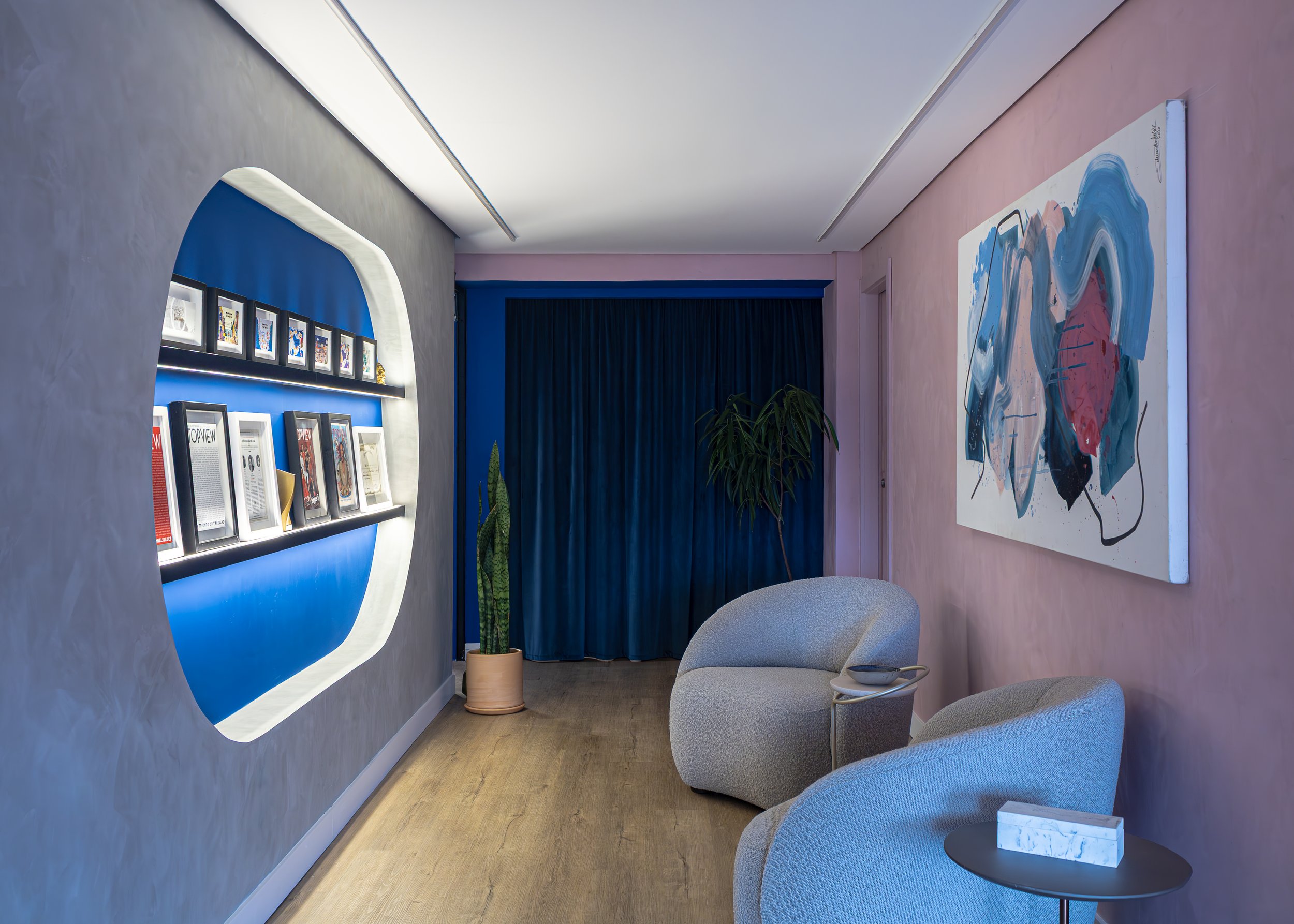 Sala de estar com duas poltronas cinzas, uma mesa de canto com uma caixa branca de papel recuperado, parede rosa com quadro de arte abstrata, cortinas azuis e duas plantas verdes ao fundo.