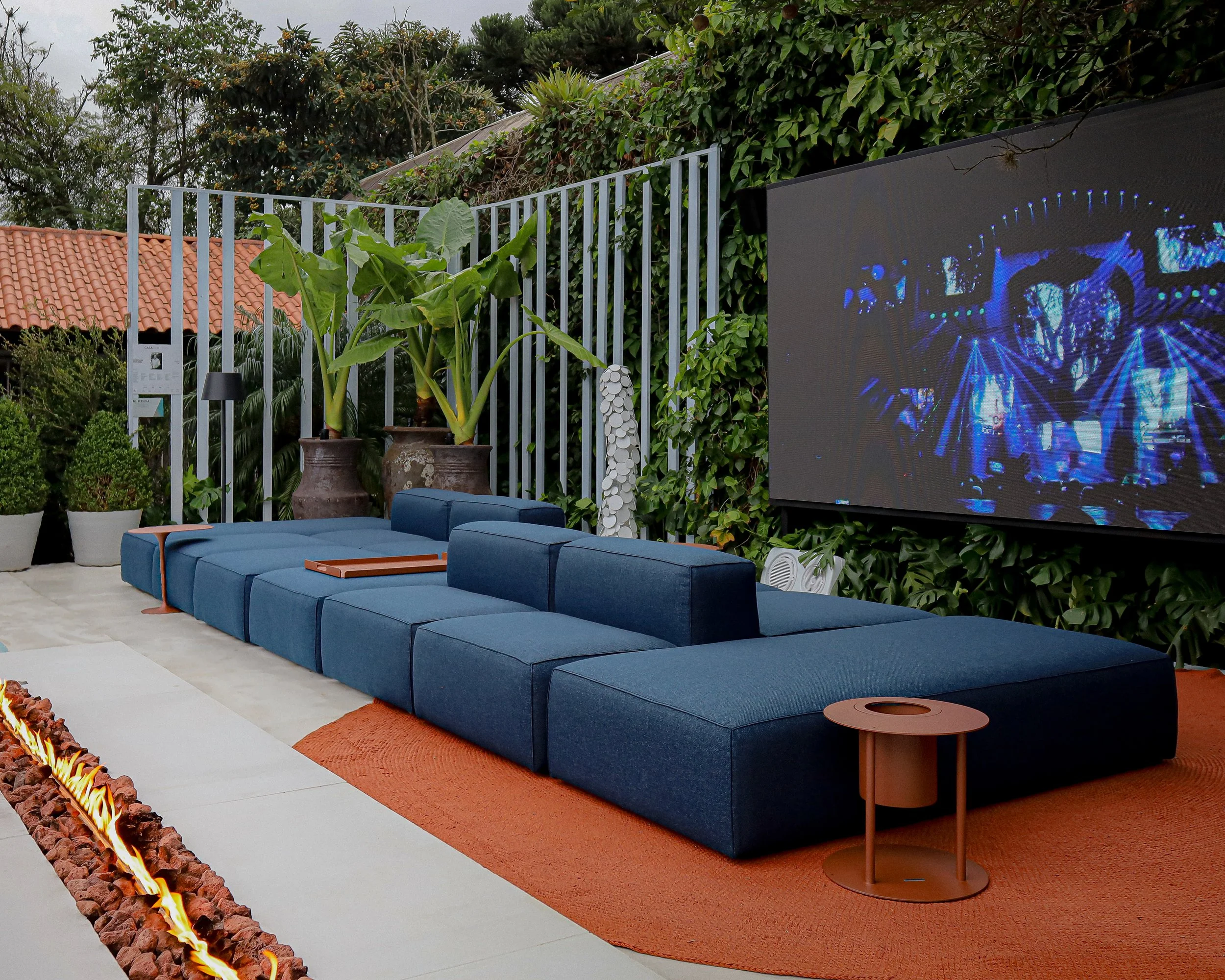 Área de convivência ao ar livre com sofá de tecido azul, mesa lateral de madeira, plantações em vasos ao fundo, tela de projeção com luzes e vegetação ao redor.