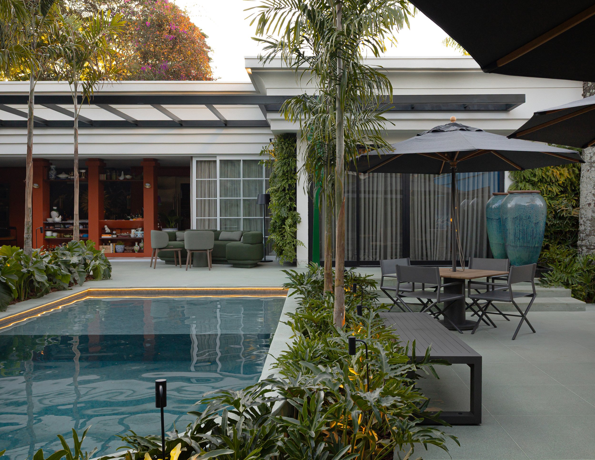 Área externa de casa com piscina, jardim, mesas com guarda-sol e sofá.