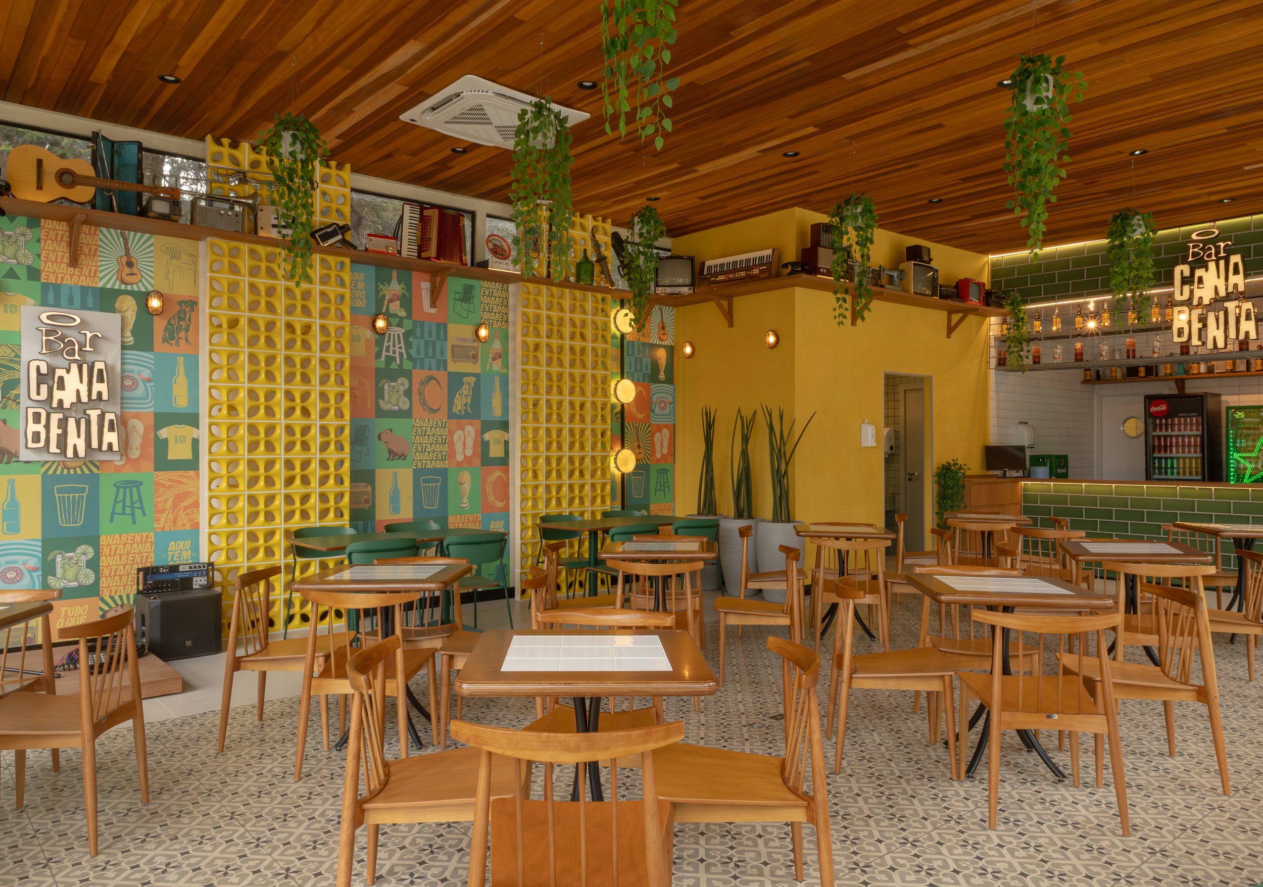 Interior de um bar com mesas de madeira, paredes decoradas com cores vibrantes, plantas penduradas no teto e uma parede com colagens de quadros e objetos retro, criando um ambiente acolhedor e descontraído.