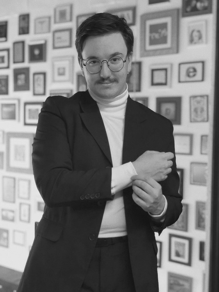 Homem usando óculos, terno preto e suéter branco, posando com braços cruzados em frente a parede com quadros decorativos.