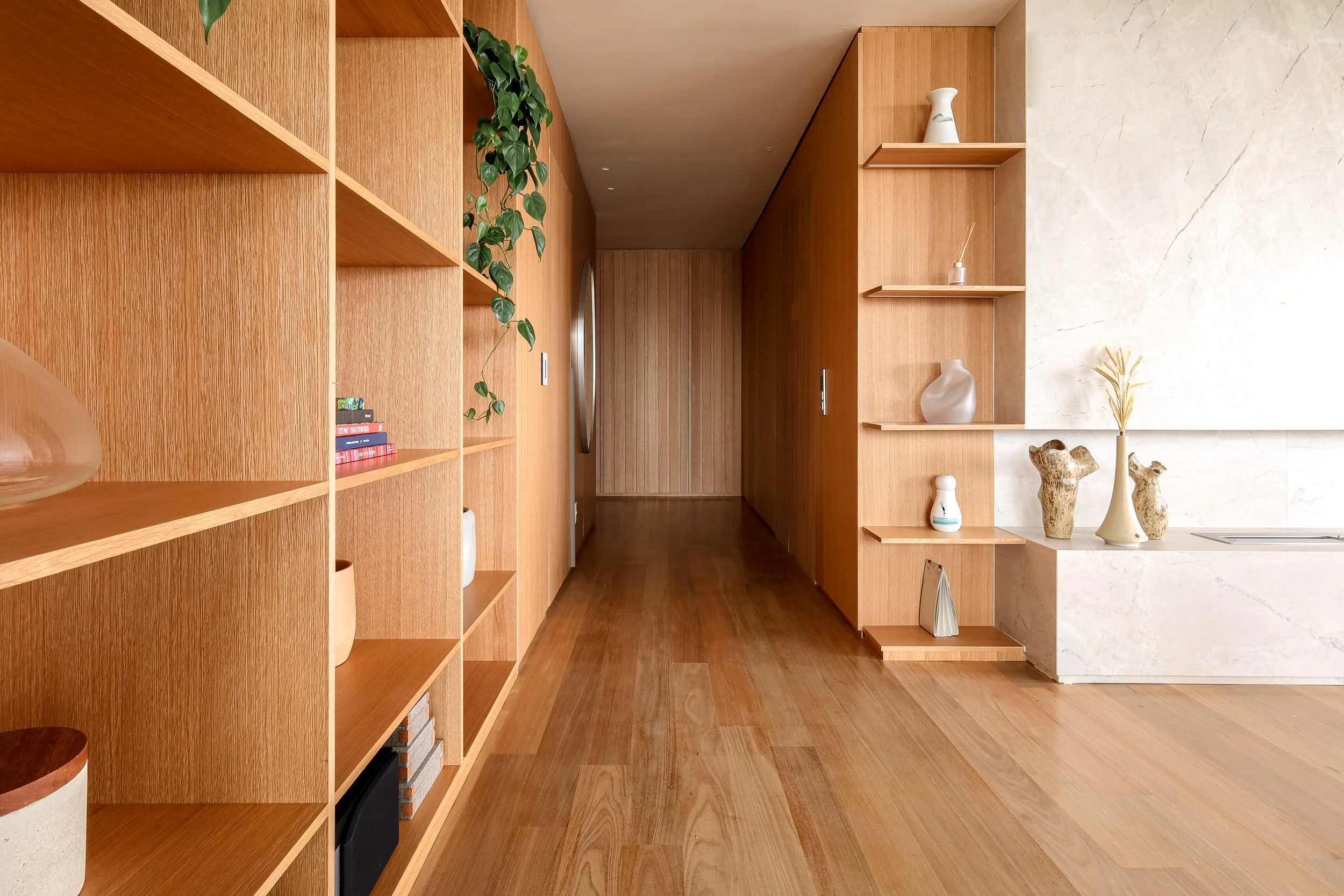 Corredor com paredes de madeira e piso de madeira, com prateleiras decorativas de ambos os lados, contendo vasos, livros e objetos decorativos, com iluminação sutil.
