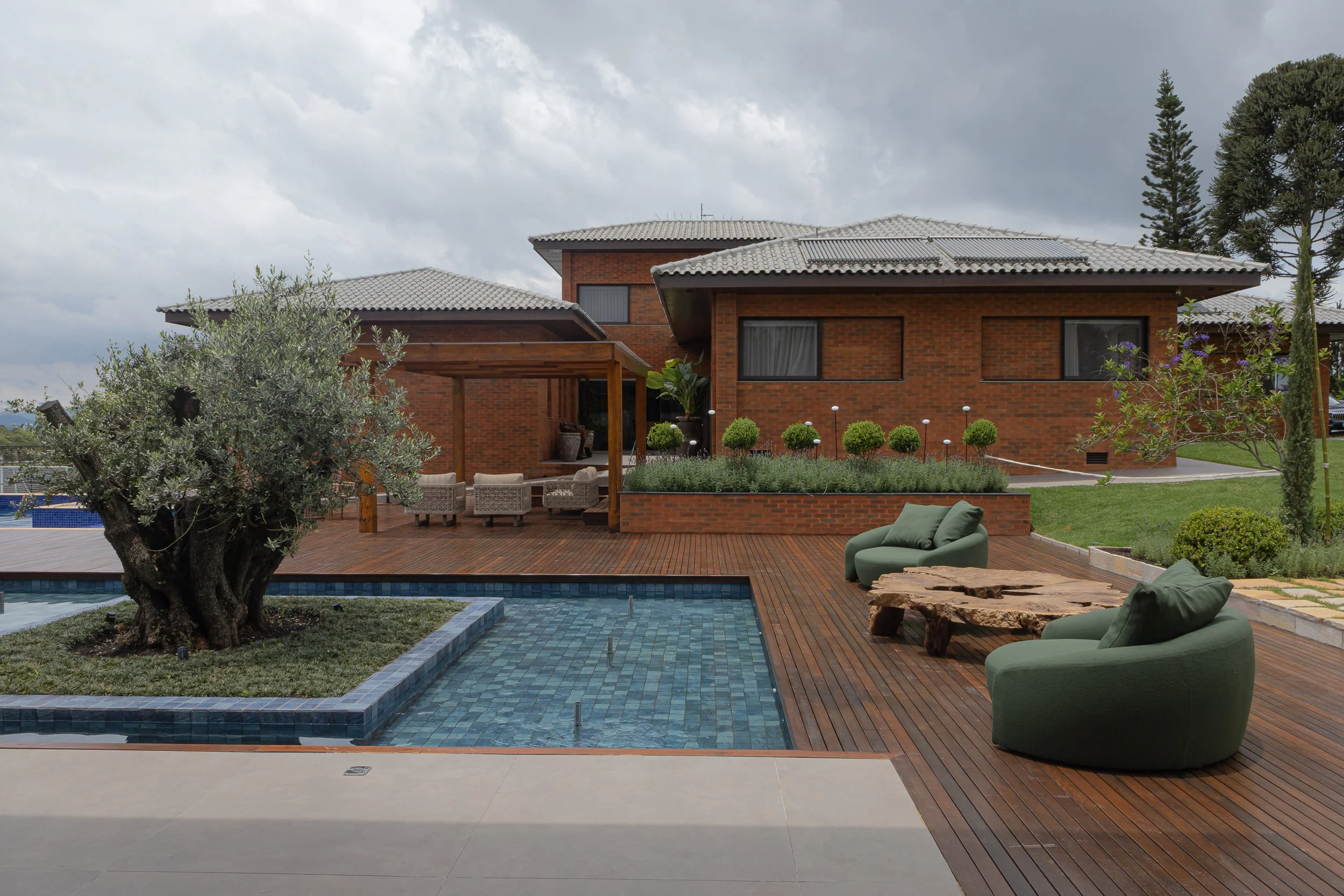 ÁREA externa de casa com piscina de azulejos azuis, árvore ao centro, sofá de canto verde, mesa de madeira e jardim com plantas e arbustos em fundo de casa de tijolos vermelhos.