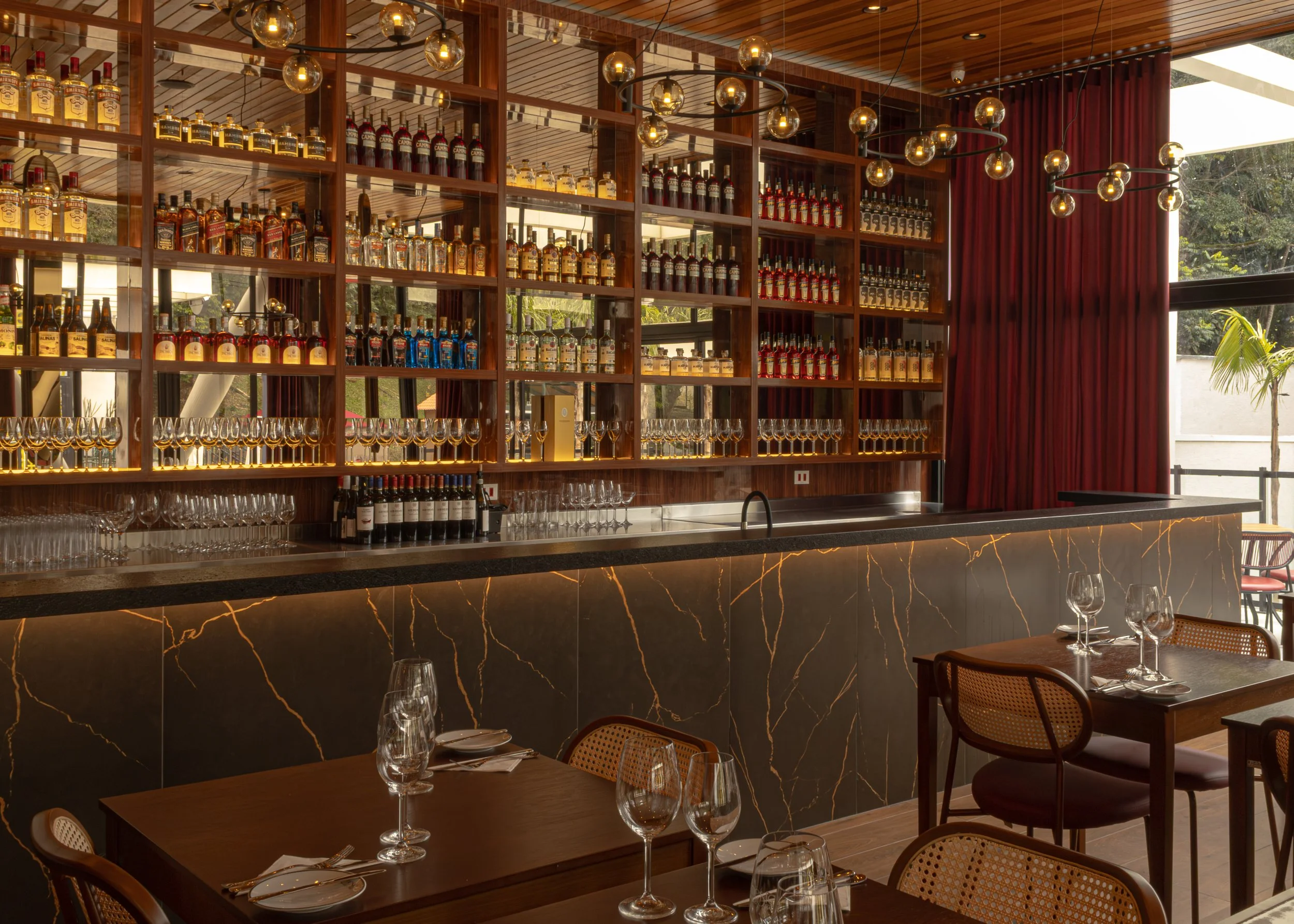 Bar moderna com prateleiras de bebidas alcoolicas, copos de vinho e taças no balcão, mesas com taças de vinho e pratos, cortinas vermelhas e iluminação contemporânea