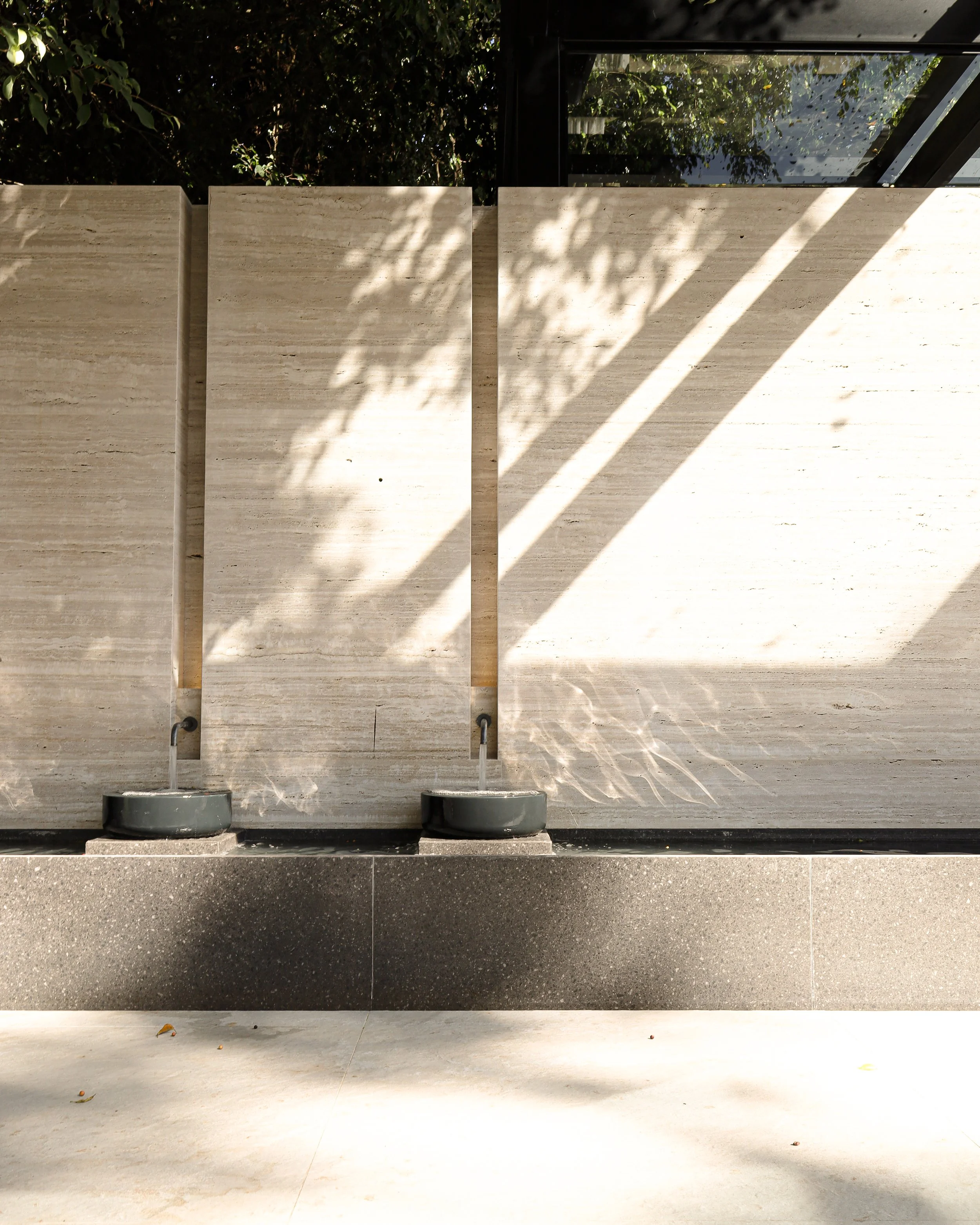 Duas fontes de água de pedra preta em uma parede de blocos de pedra clara, com sombras de árvores projetadas nela. Há uma cobertura de vidro no canto superior direito.