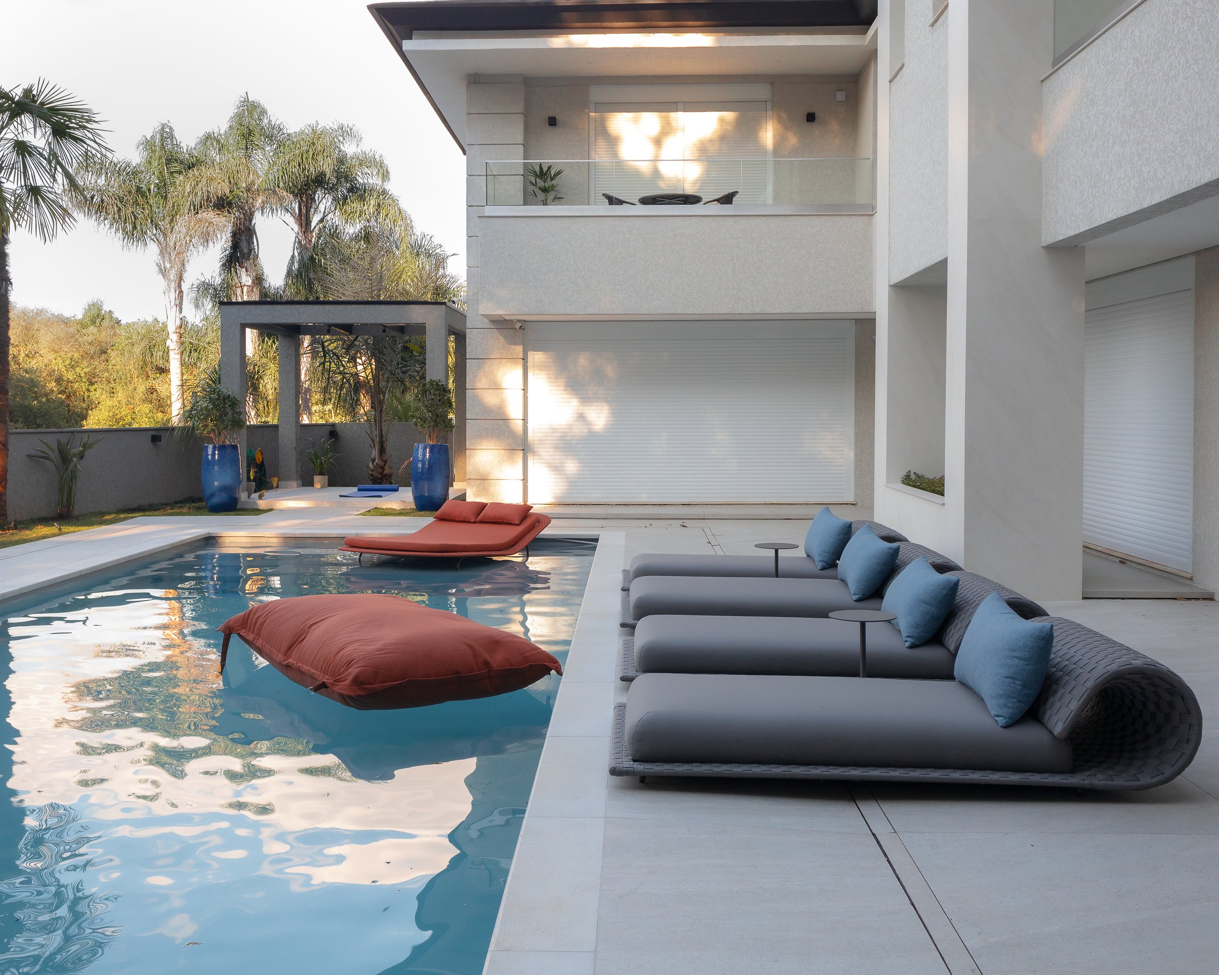 Área de lazer com piscina, espreguiçadeiras, almofadas coloridas, plantas em vasos e varanda com móveis na casa moderna