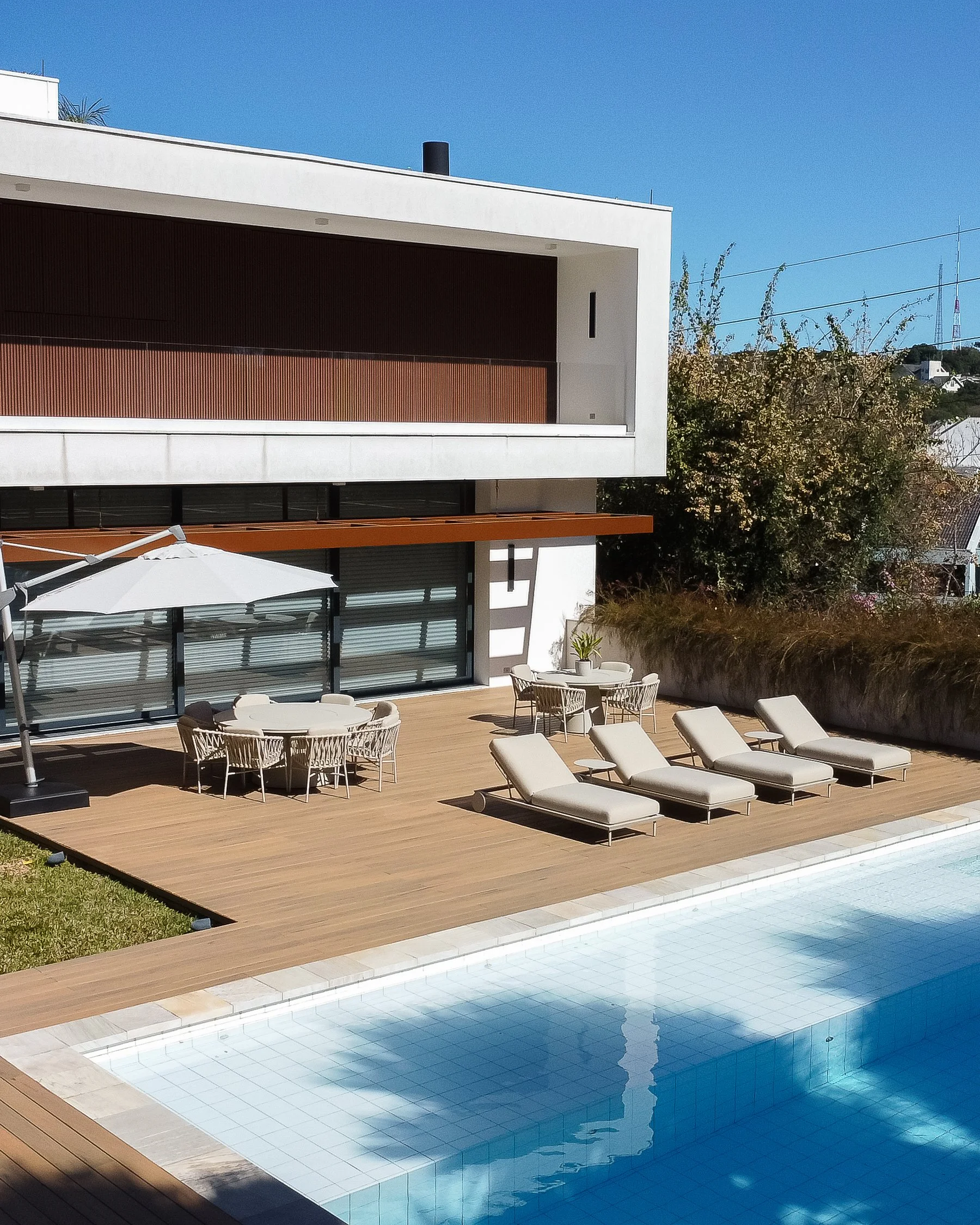 Piscina e área de lazer com espreguiçadeiras, mesas, cadeiras e guarda-sol, em frente a uma casa moderna.