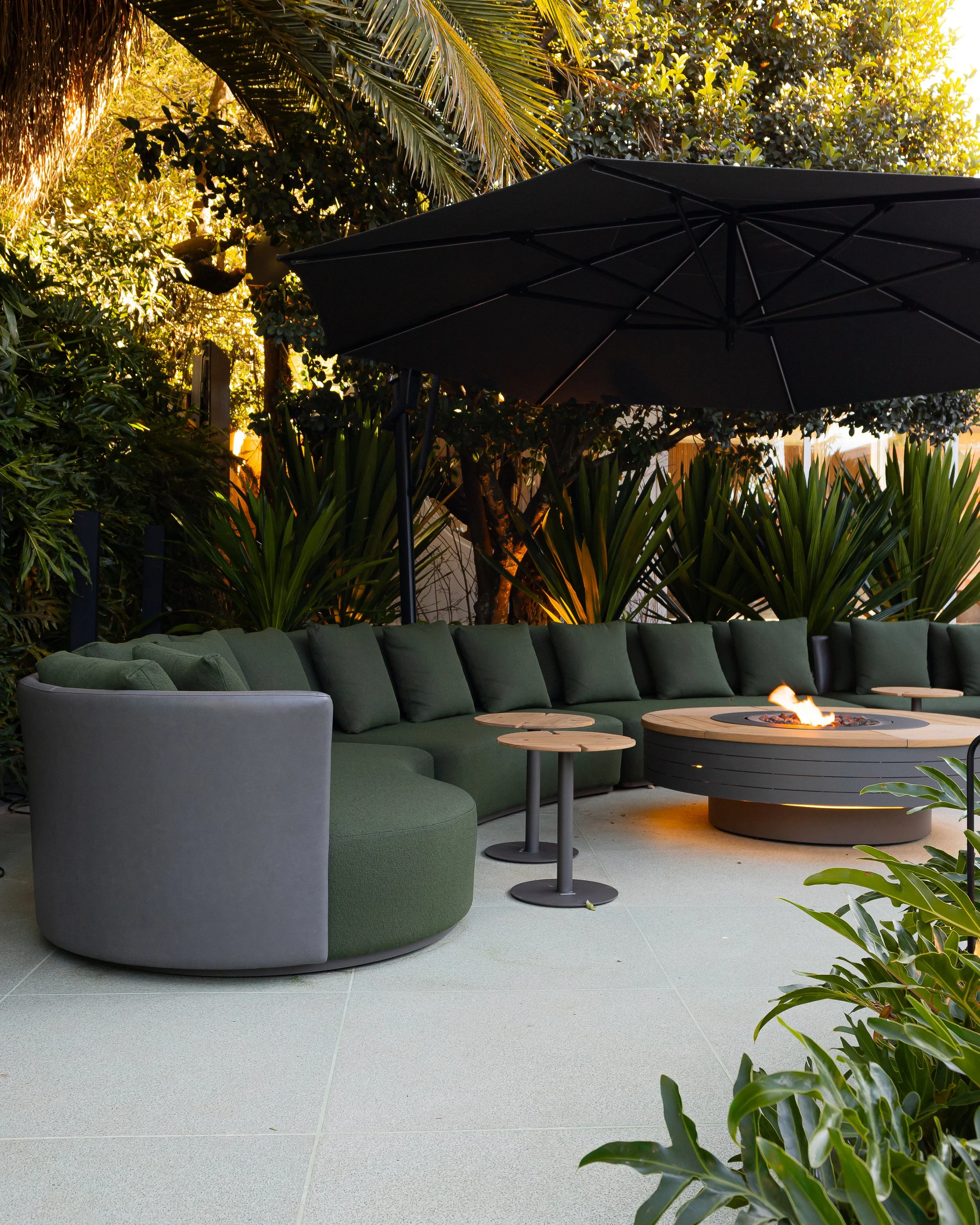 Área de convivência ao ar livre com sofá circular grande de almofadas verdes, espreguiçadeira cinza. Mesa de centro com fogo artificial, cobertura de guarda-sol preto e plantas ao redor.