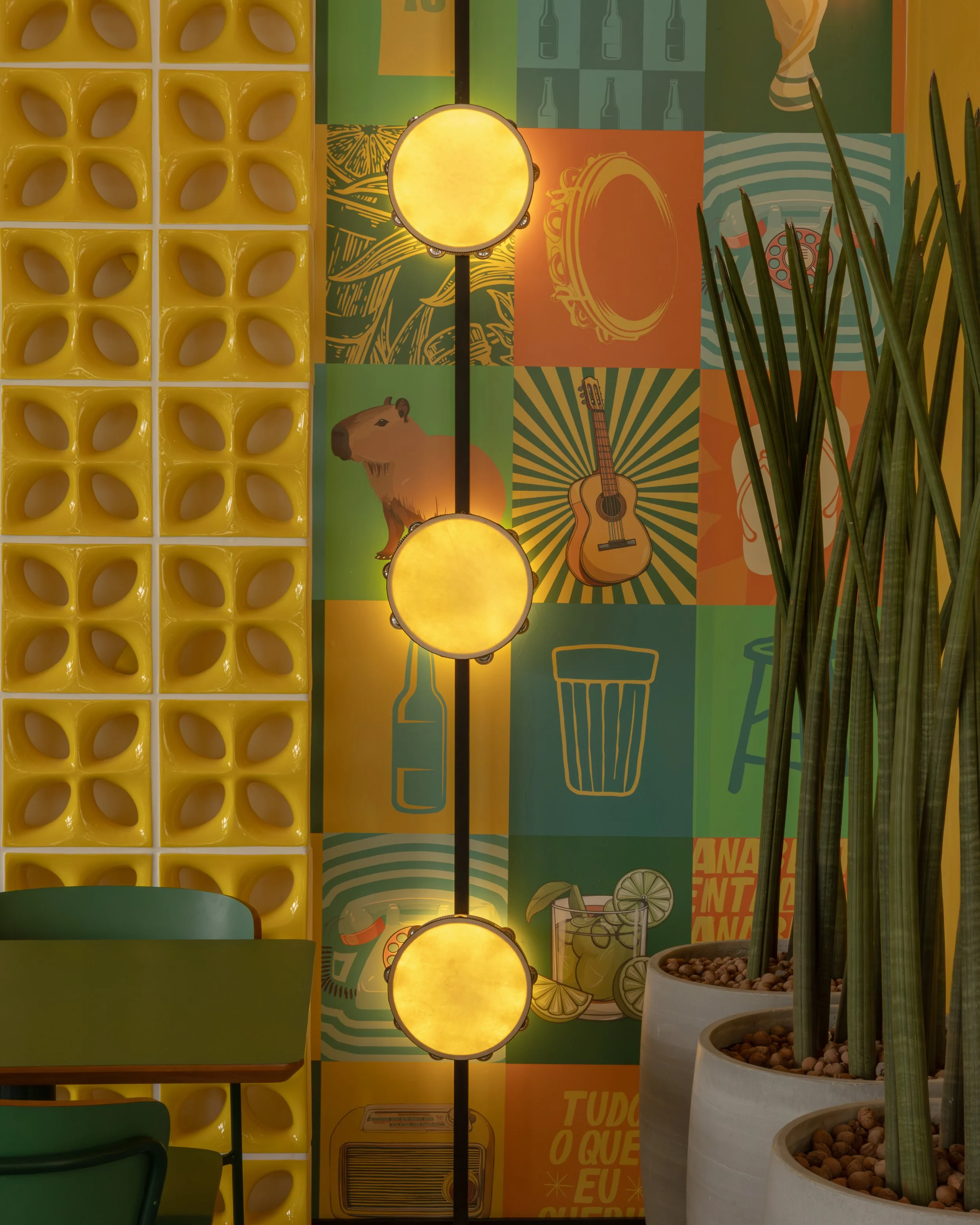 Parede decorada com ilustrações coloridas de objetos musicais, bebidas e animais, ao lado de uma parede amarela com detalhes de buracos em forma de folha, e um vaso grande com plantas altas.