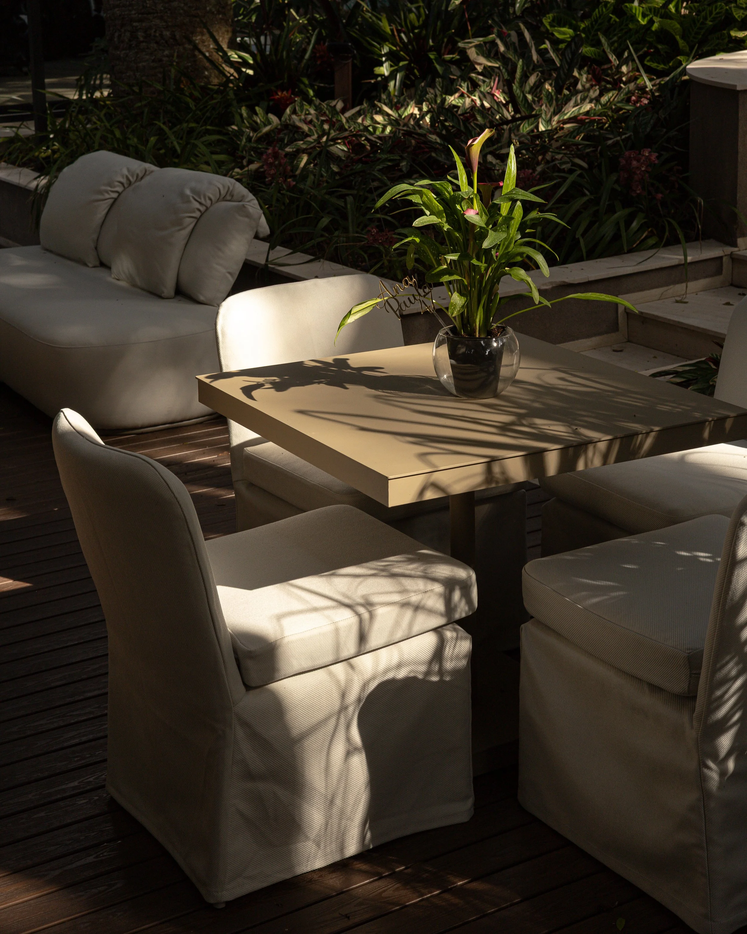 Mesa de jantar com uma planta de flores no centro, cercada por cadeiras com estofado bege, em ambiente com luz solar e vegetação ao fundo.