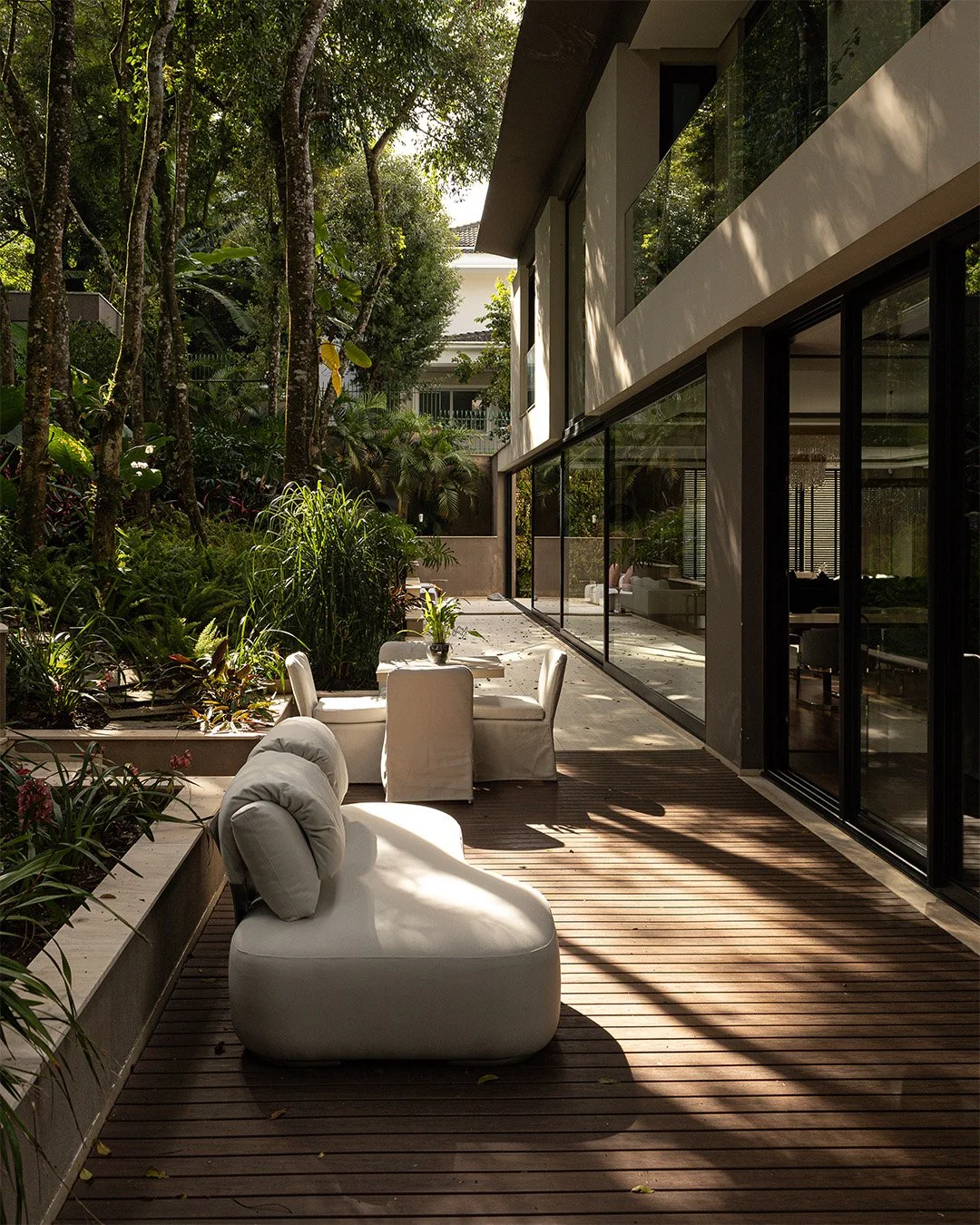 Varanda moderna com móveis brancos e plantas em uma residência, cercada por árvores verdes e vegetação, com janelas de vidro.